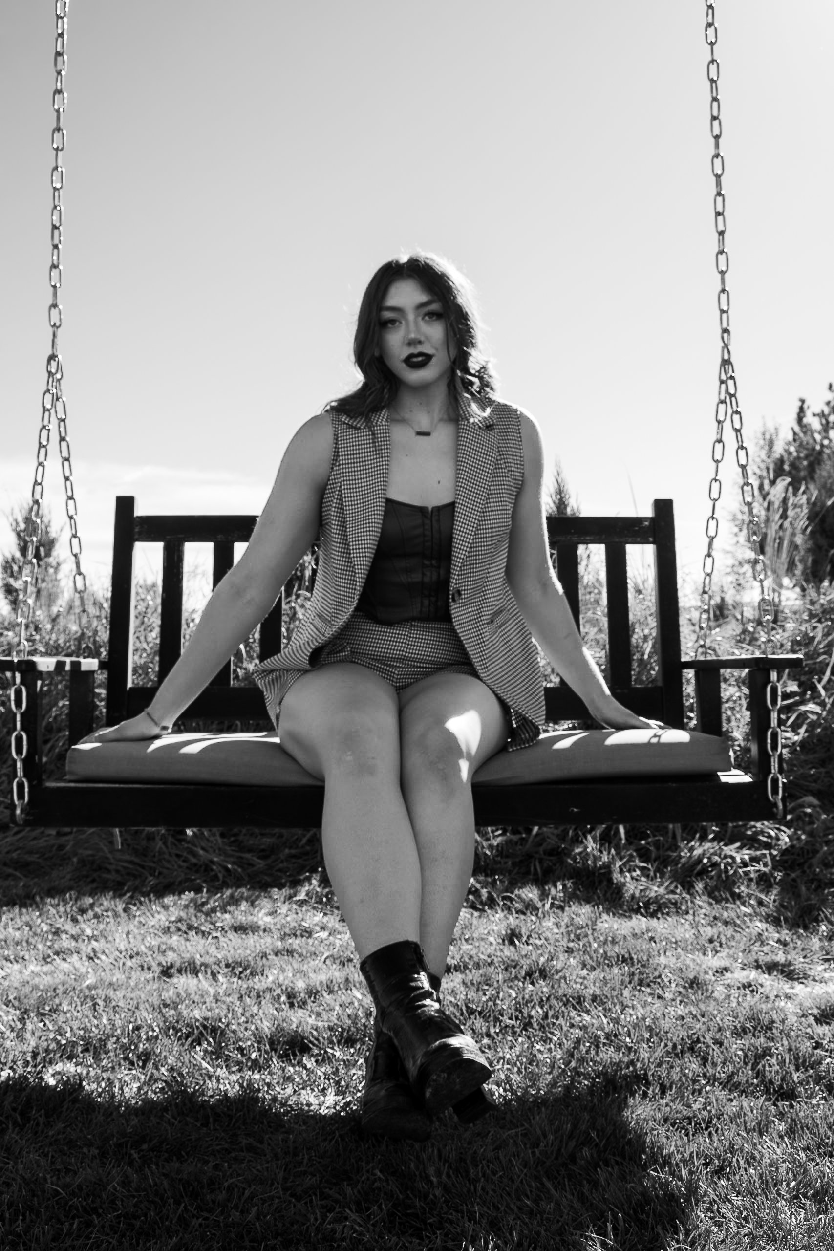 A woman sitting on a swing outdoors during daytime, dressed in a checkered vest, shorts, and boots, with dark lipstick and styled hair.