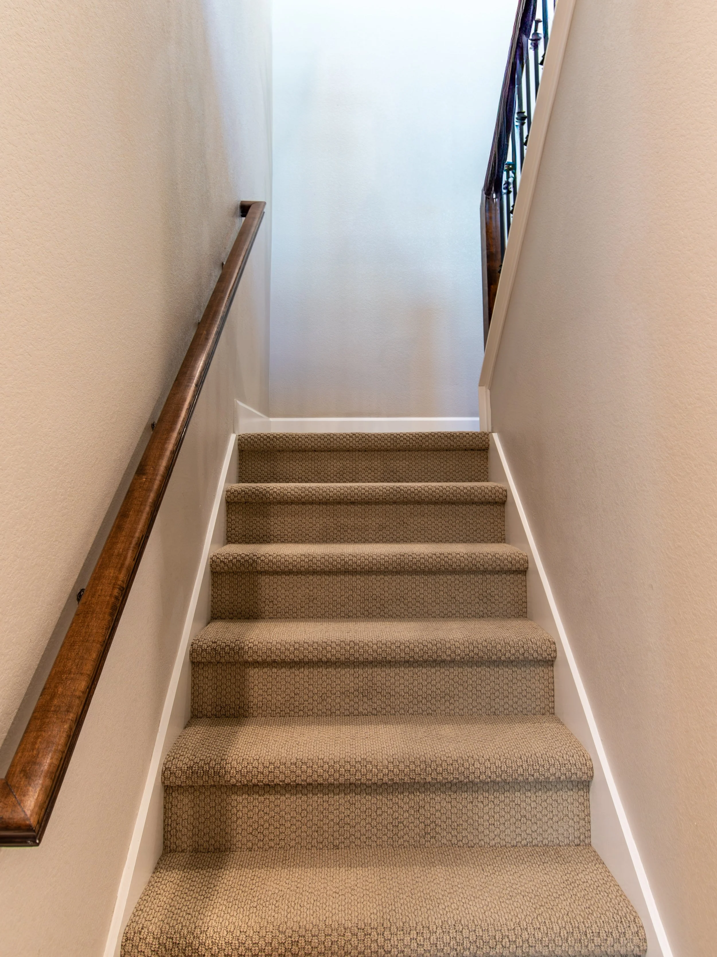 Carpeted staircase with wooden handrail on the left and a wall on the right, leading up to a brightly lit upper floor.