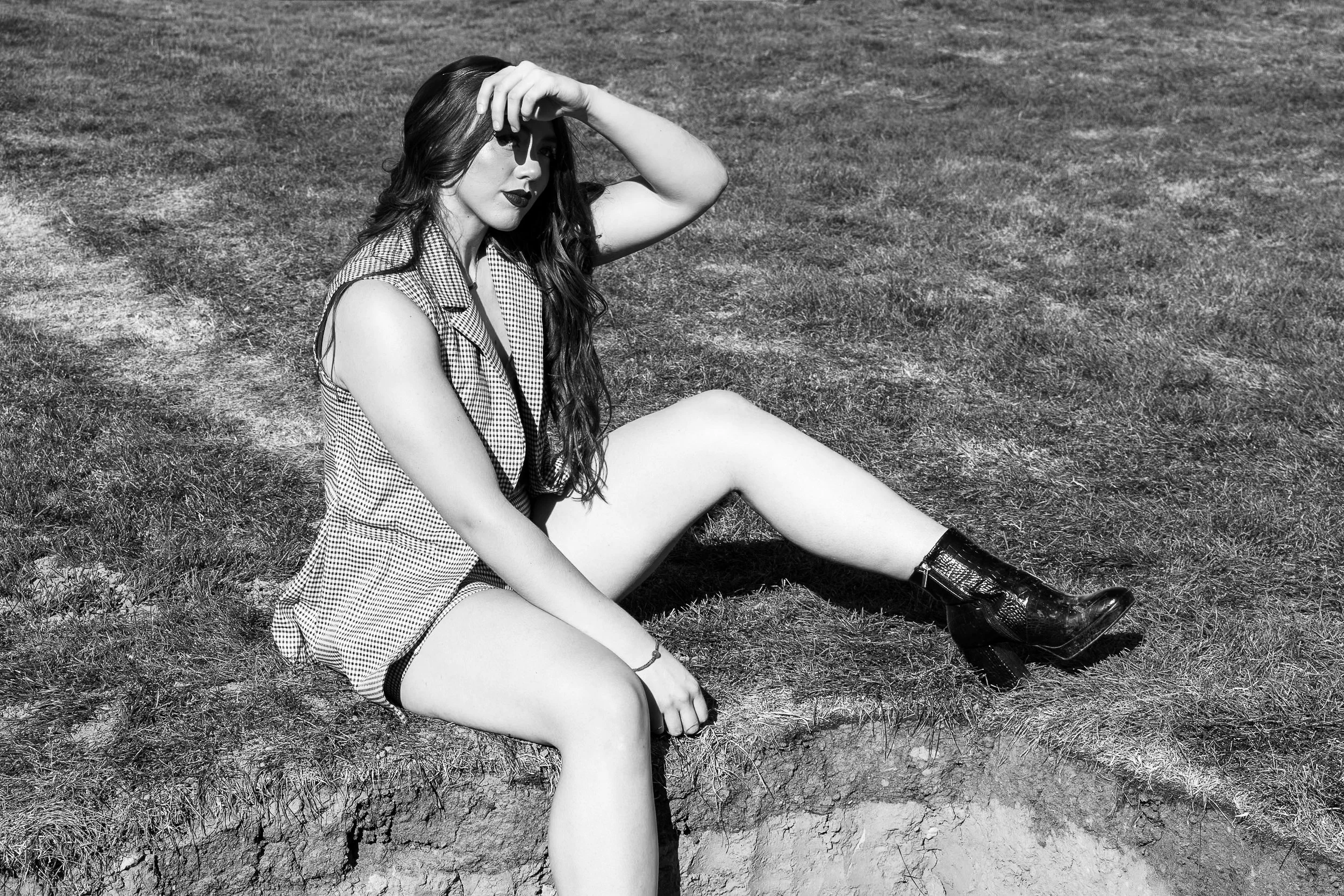 A woman with long hair sits on the grass with her legs extended, wearing a checkered sleeveless shirt and boots, shading her eyes from the sun.