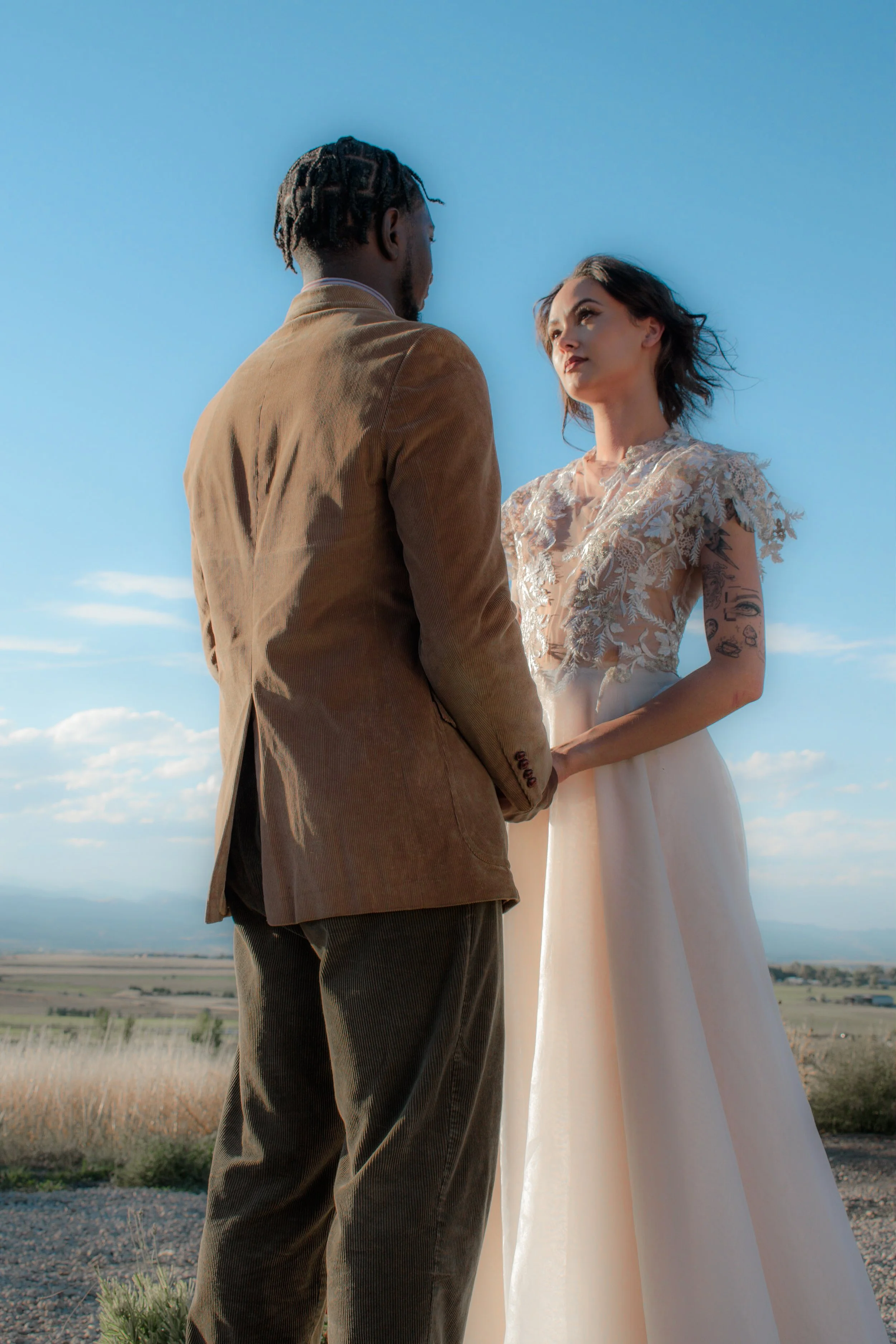 A couple holding hands outdoors against a blue sky, with the man wearing a brown jacket and the woman in a wedding dress with lace details.