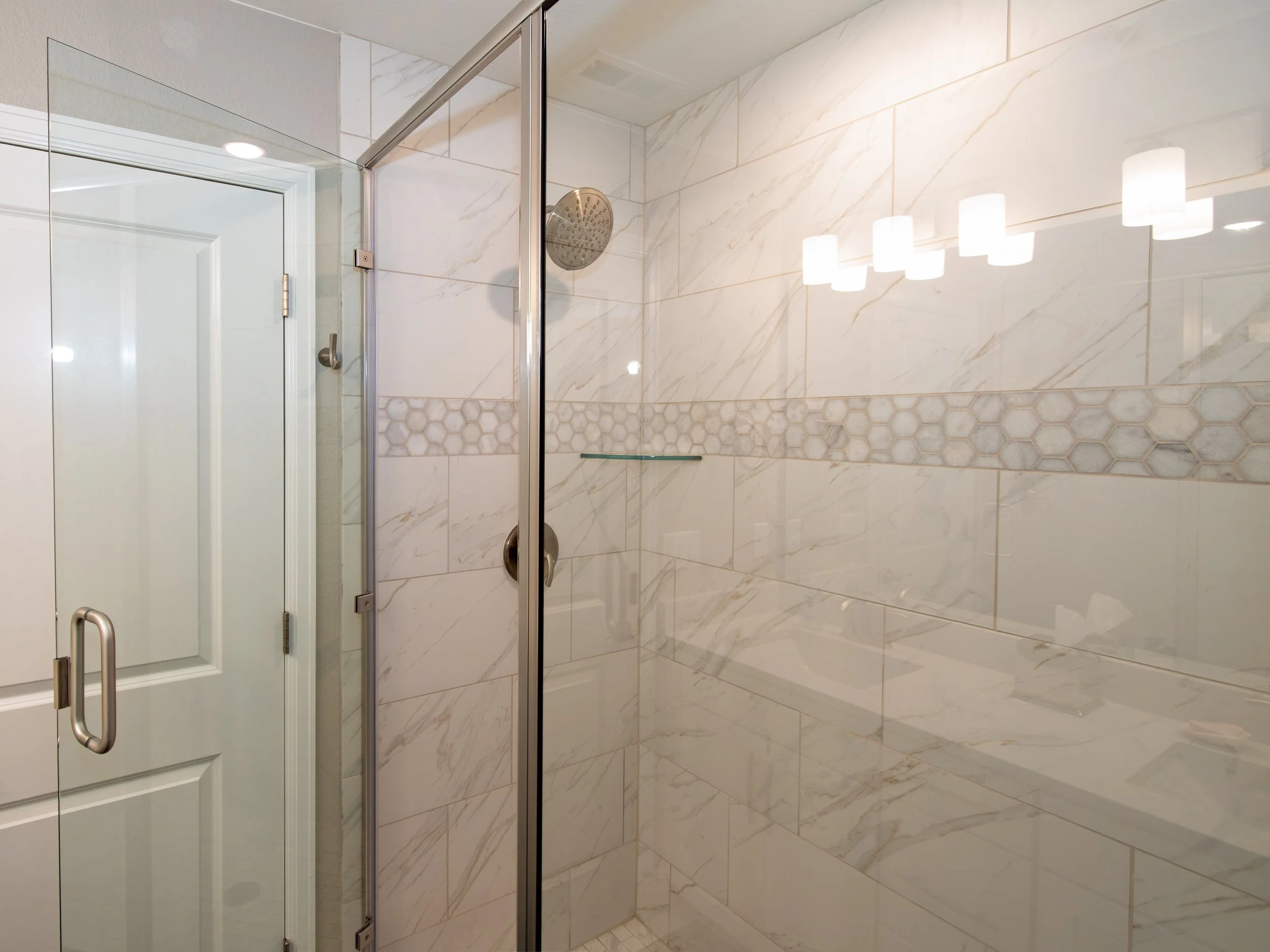 Inside a modern bathroom shower with large marble tiles, a glass door, a rain shower head, and a decorative tile strip.