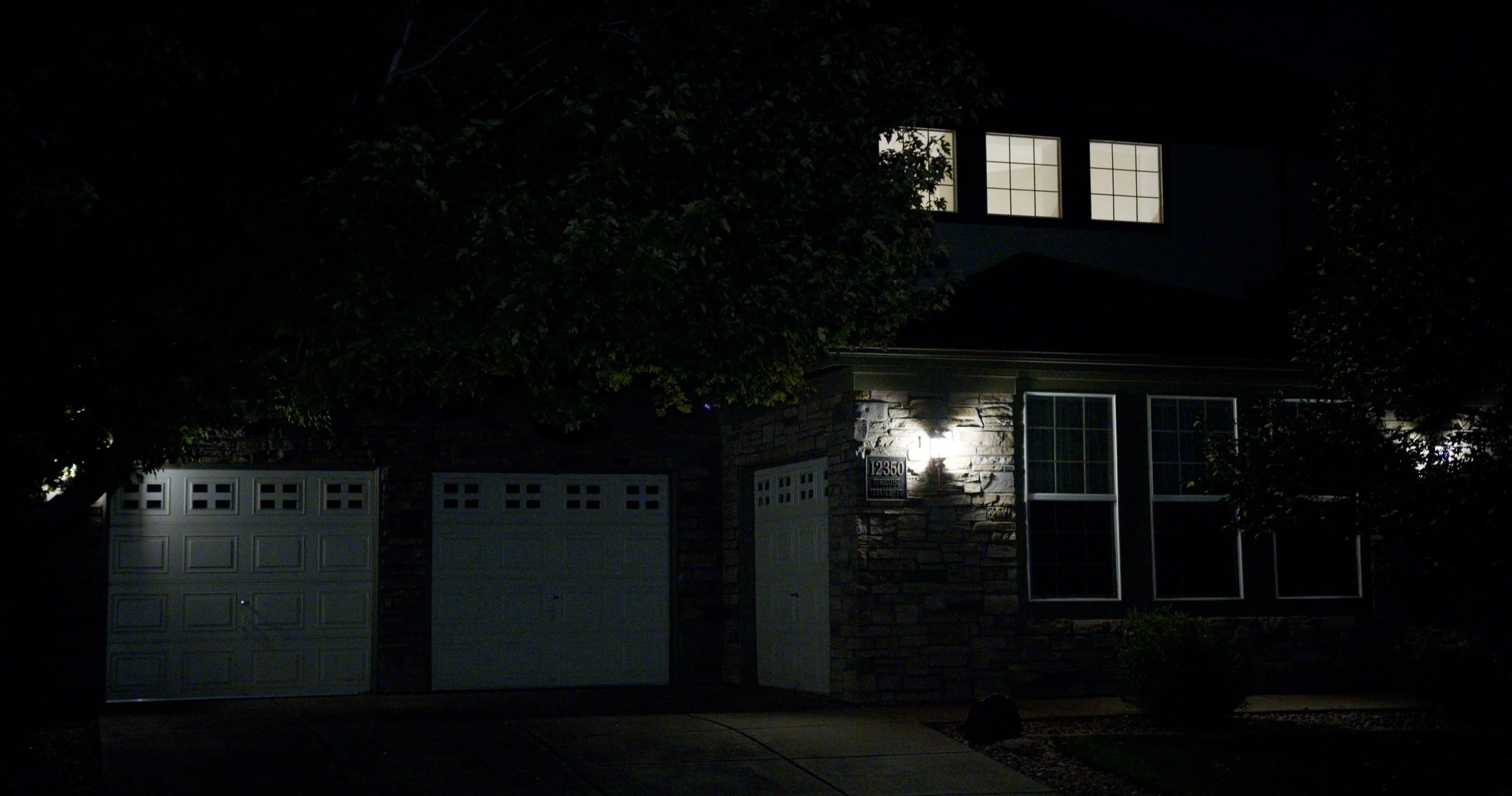A house at night with illuminated window and exterior lights, stone exterior wall, attached garage with closed doors, and trees partially obscuring the view.