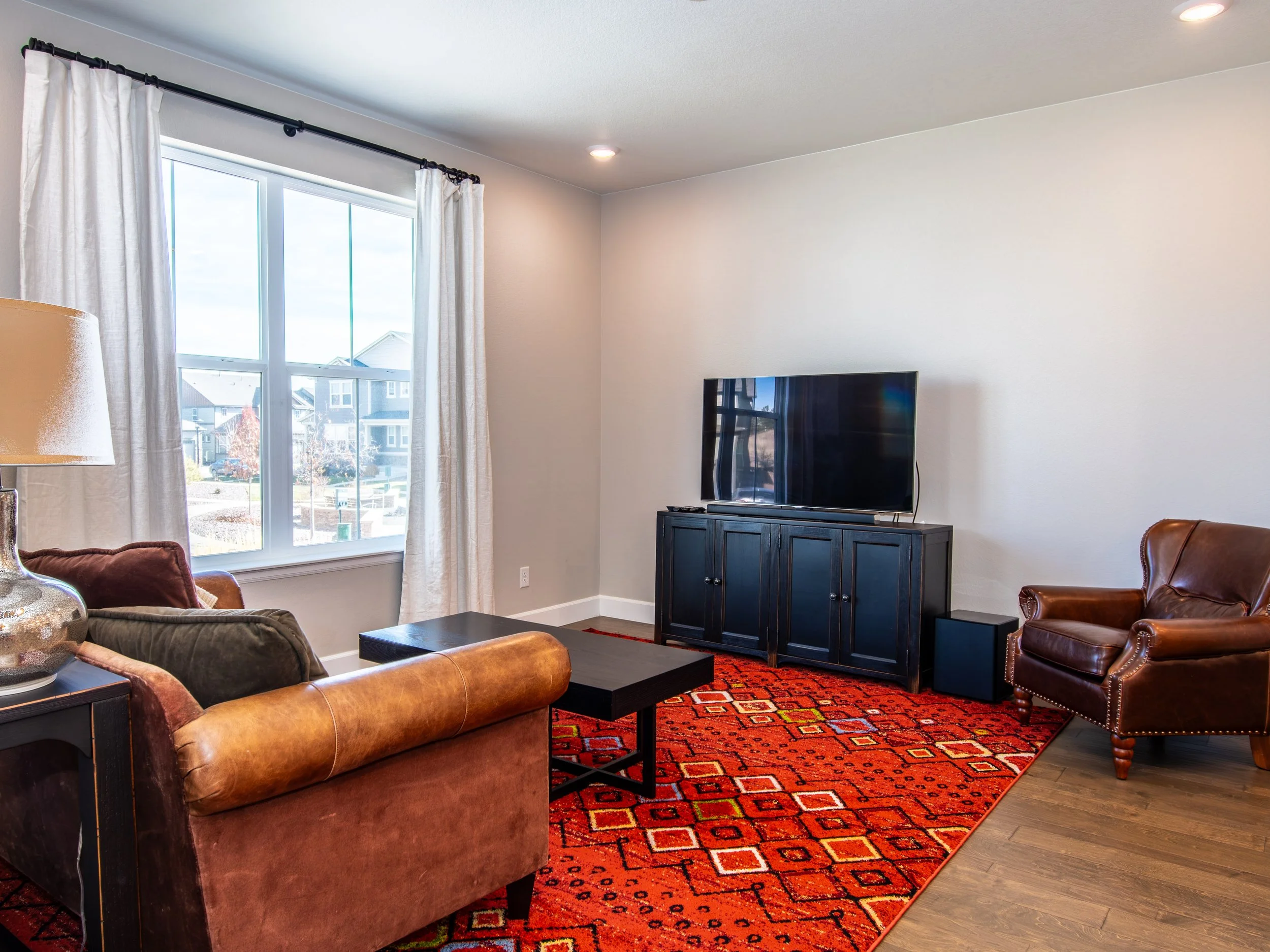 Living room with large window, beige walls, leather armchair, sofa, black coffee table, red patterned area rug, and flat-screen TV on a black TV stand.
