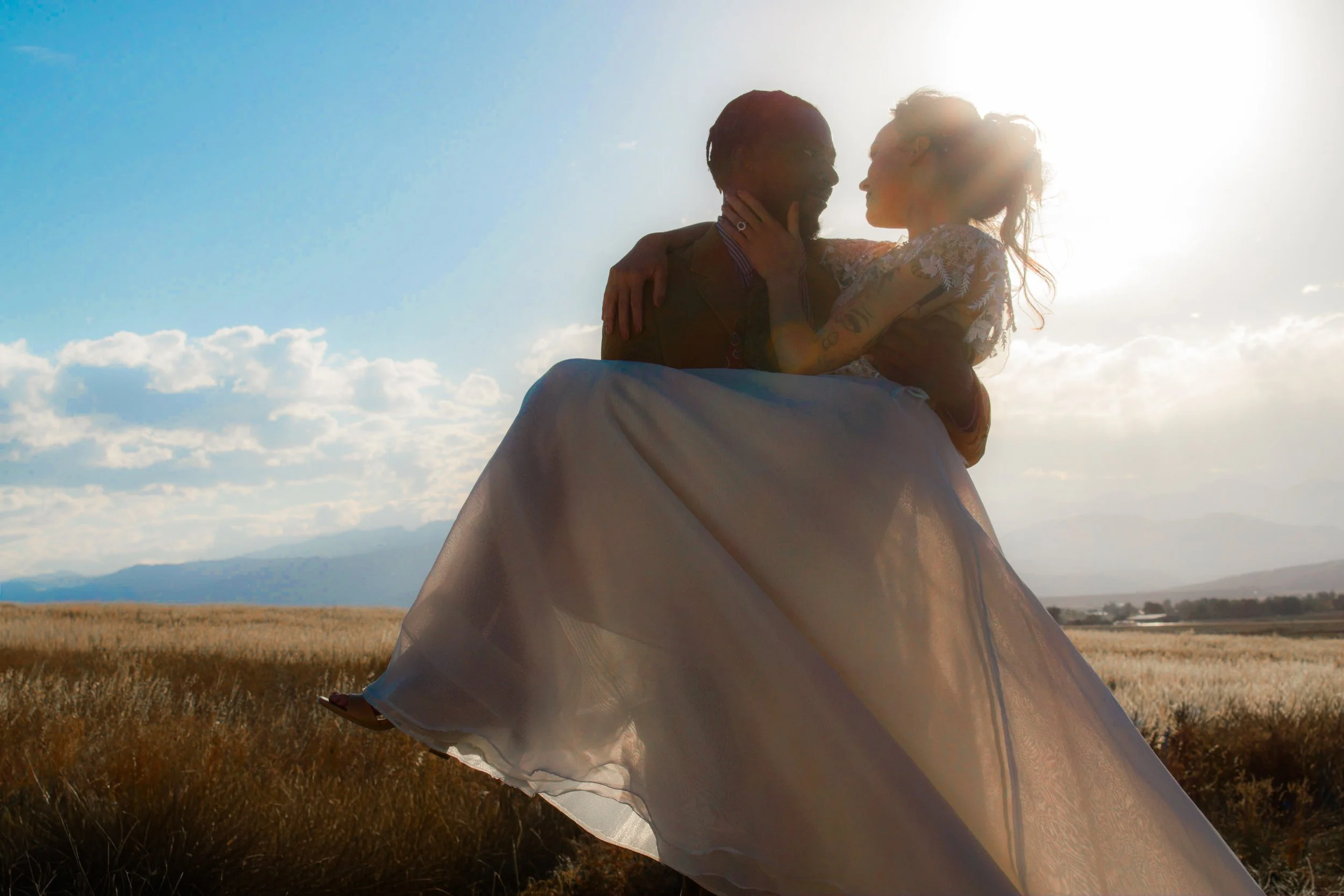 A man carrying a woman in a flowing dress in a field with mountains in the background, backlit by the sun.