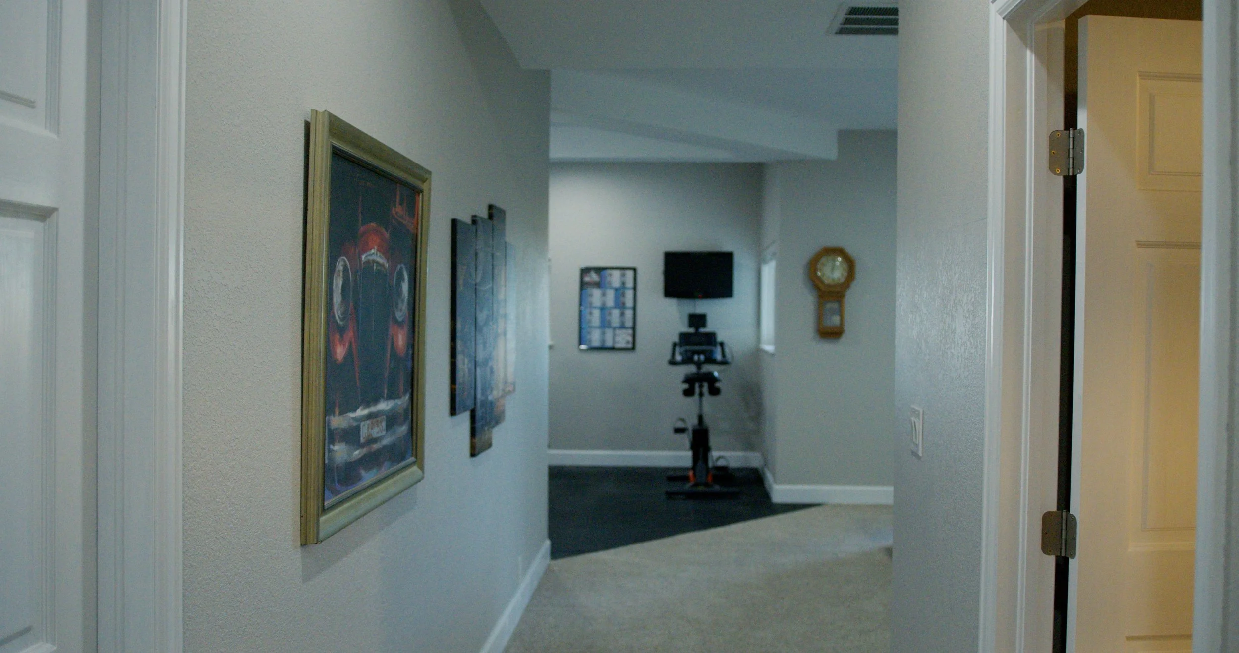 Hallway with framed pictures on the wall and a wooden clock, leading to a room with exercise equipment and a television mounted on the wall.