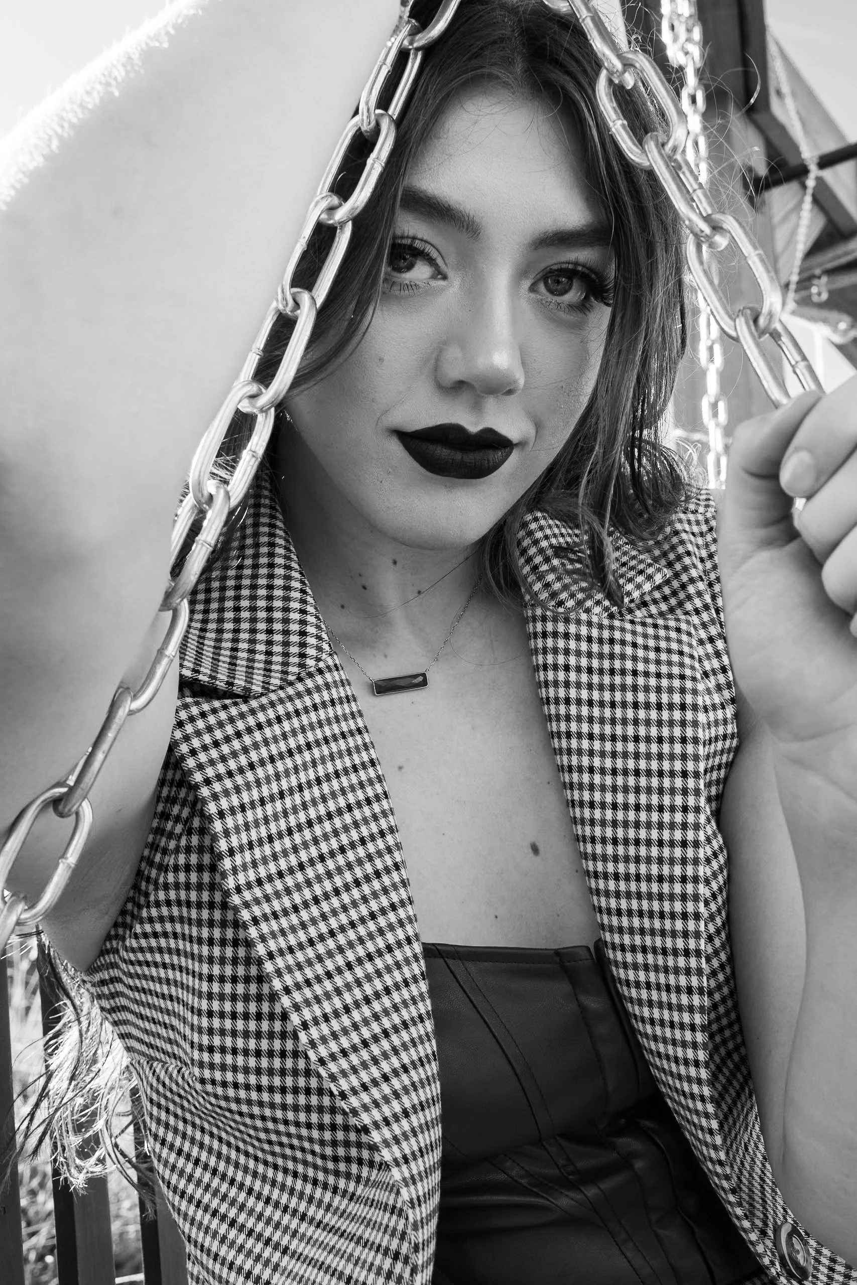 A woman with dark lipstick and wavy hair holding a swing chain, wearing a checkered jacket and gothic-style clothing, outdoors.