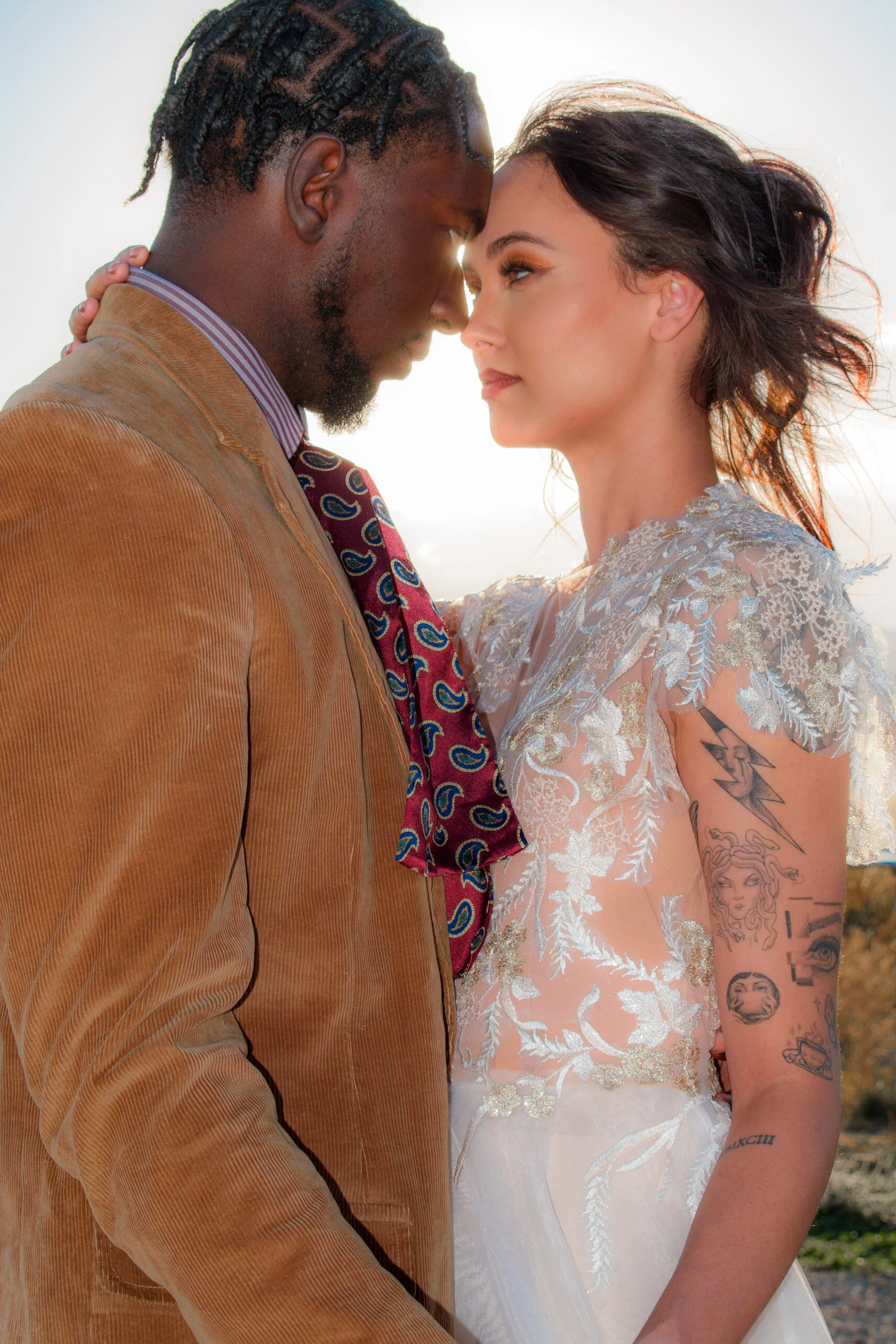 A couple stands close with foreheads touching outdoors, the man wearing a brown corduroy jacket and striped shirt, the woman in a lace wedding dress with tattoos on her arm, both gazing affectionately at each other.