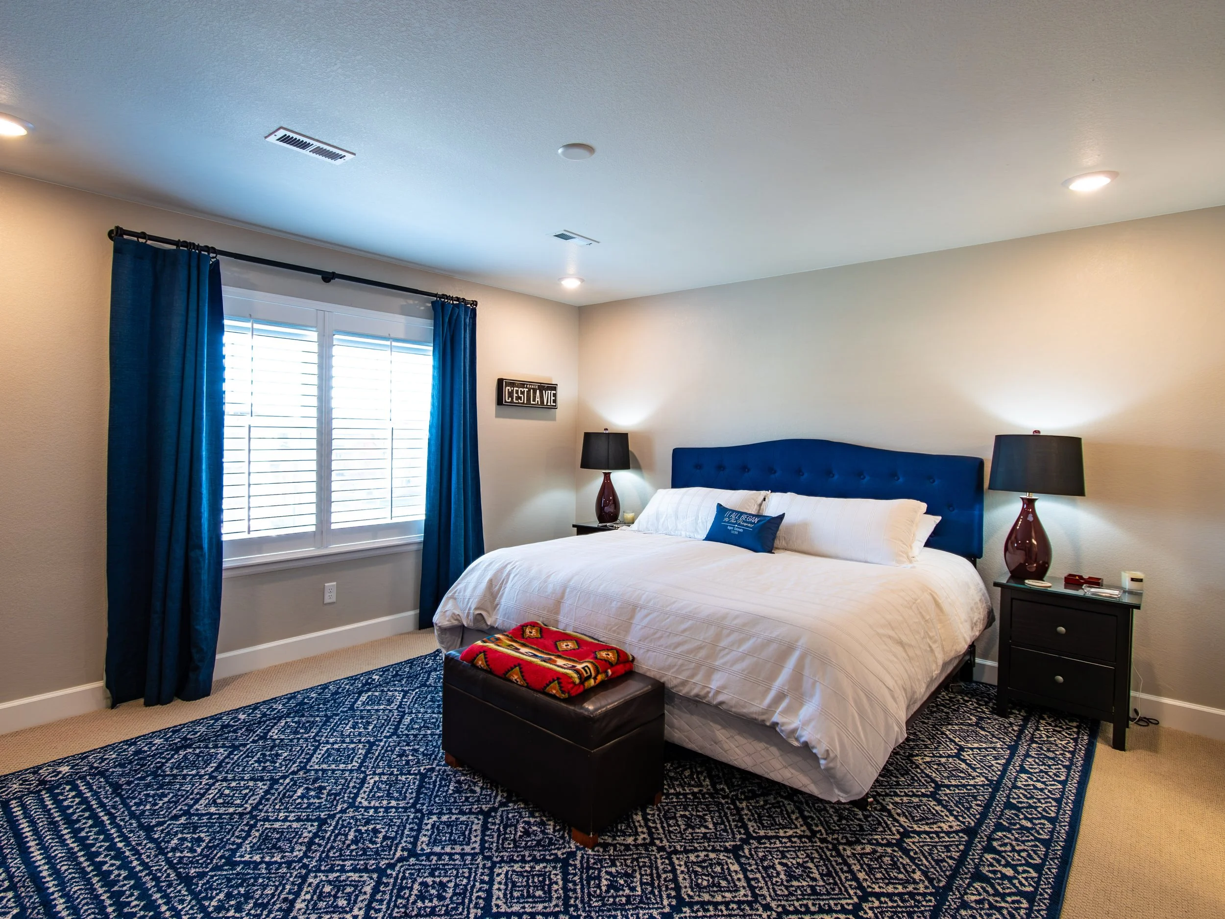 A bedroom with a window dress with blue curtains and blinds, a bed with a blue upholstered headboard, white bedding, and dark nightstands with lamps, a patterned rug, and a wall sign that says "C'est la vie."