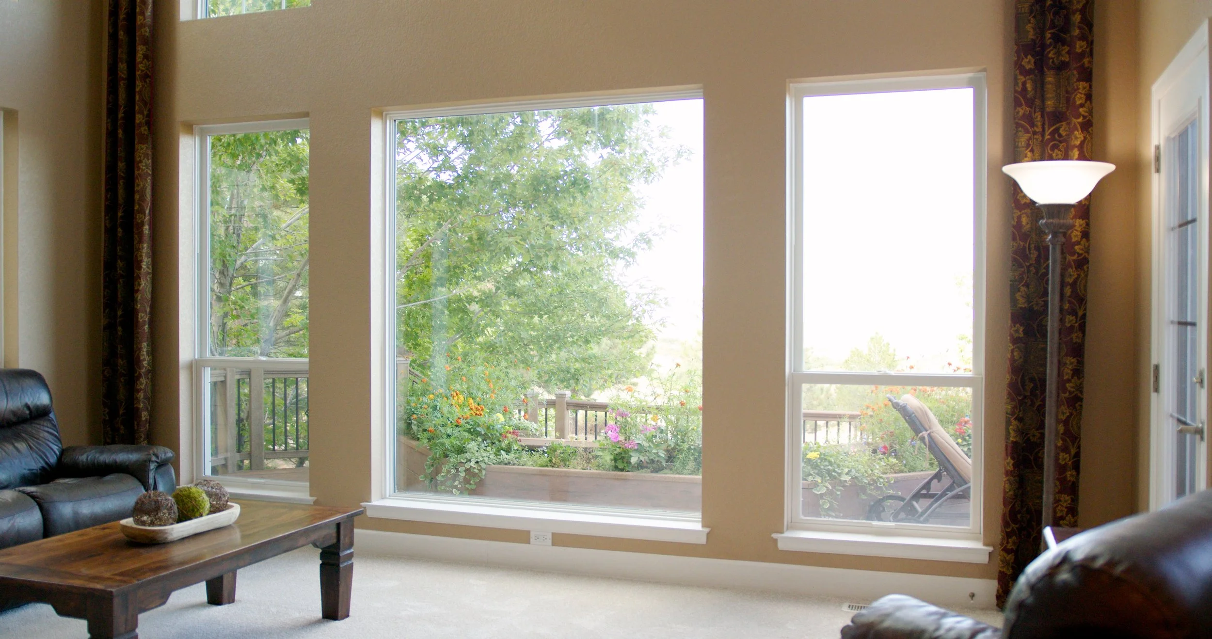Living room with large windows overlooking a garden with trees and flowers, a black leather couch, a wooden coffee table with decorative objects, and a tall floor lamp, with curtains on the sides.