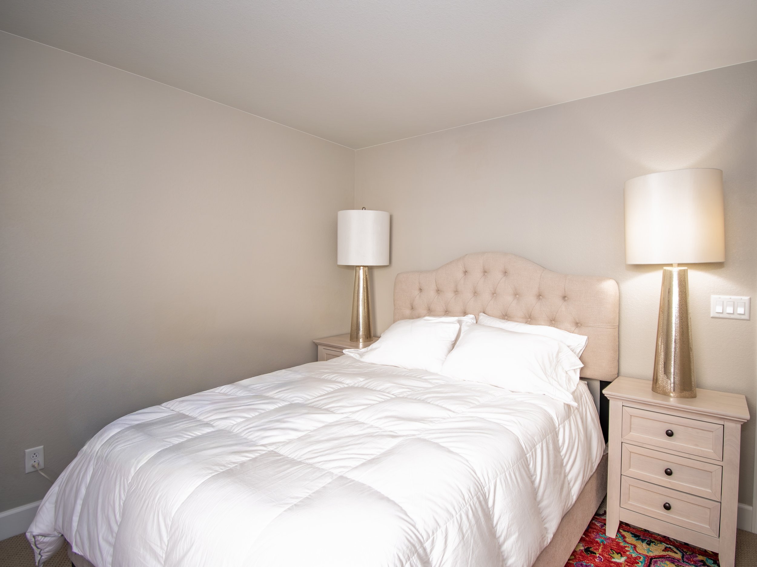 A neatly made bed with white sheets, an upholstered beige headboard, two bedside tables, and two tall lamps with white shades in a simple, clean bedroom with beige walls.
