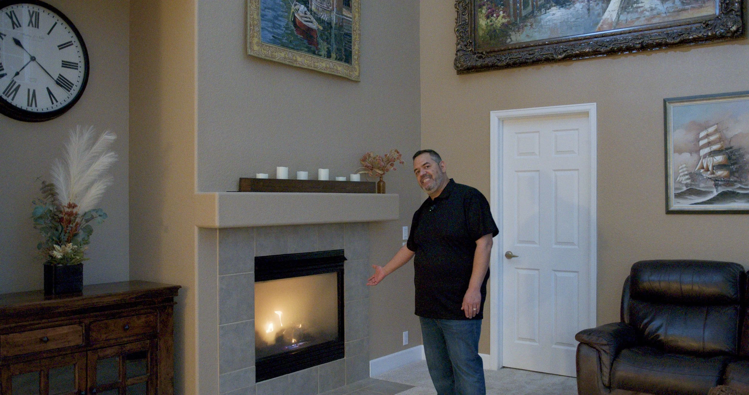 A man standing in a living room near a fireplace, pointing at it and smiling. The room has beige walls, framed artwork, a black leather chair, and a wooden sideboard with a flower arrangement. A large clock is on the wall to the left.