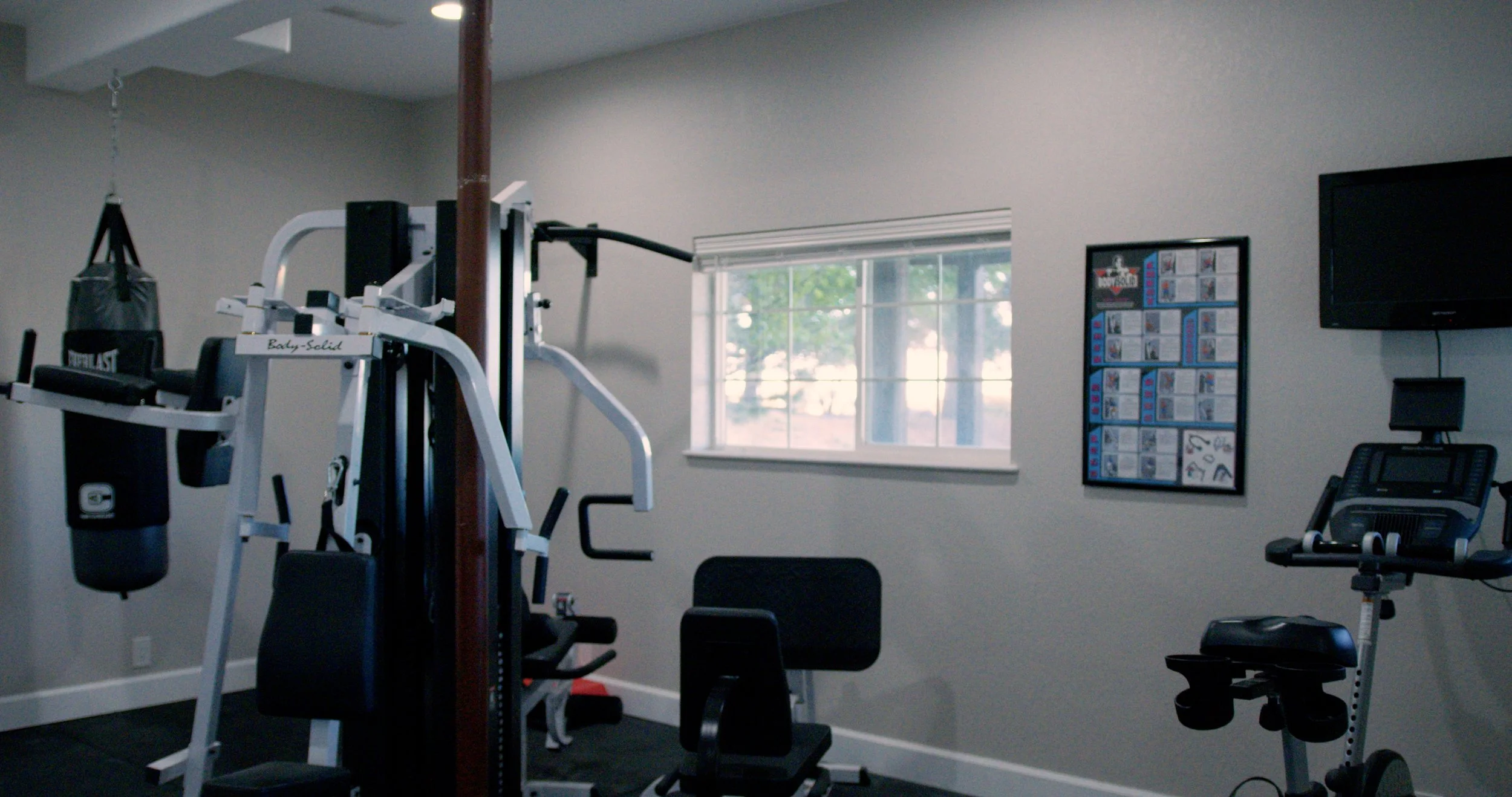 Home gym with exercise equipment including a punching bag, weight machine, stationary bike, and a wall-mounted television.