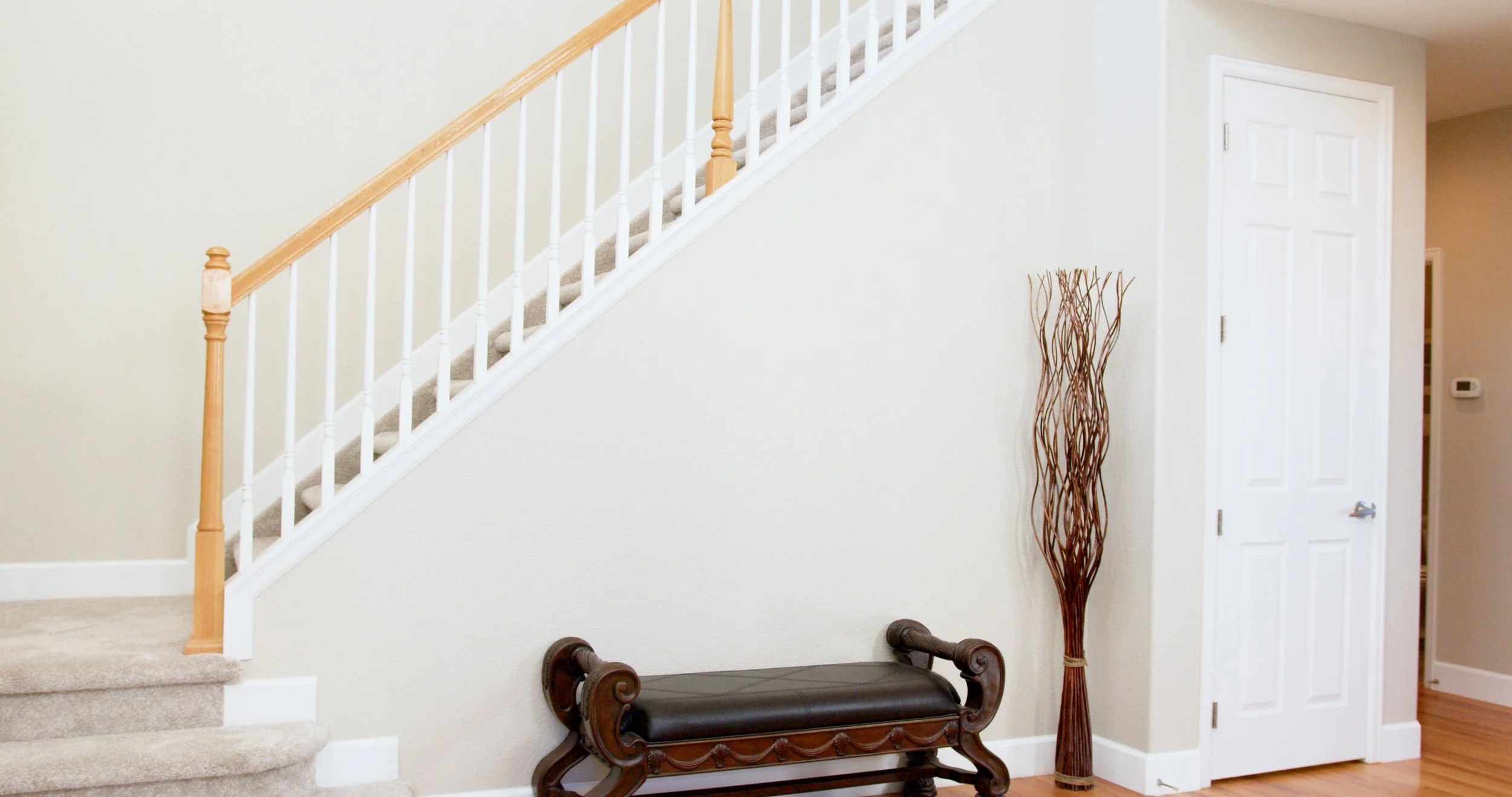 A staircase with wooden handrail and carpeted steps, a wooden bench with carved scroll arms and dark cushion, a tall decorative branch vase, and a closed white door in an interior room.