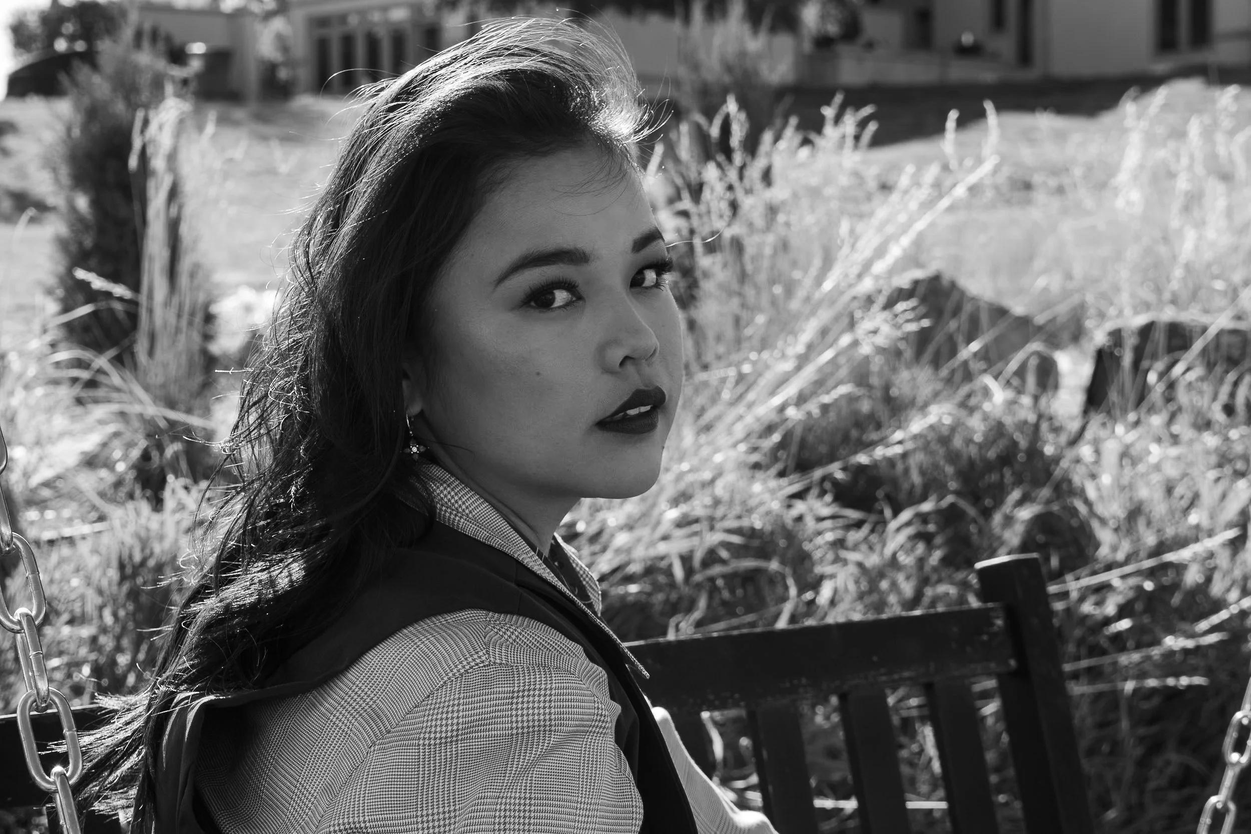 A young woman with dark hair and makeup sitting on a park bench, looking at the camera, with plants and a building in the background.