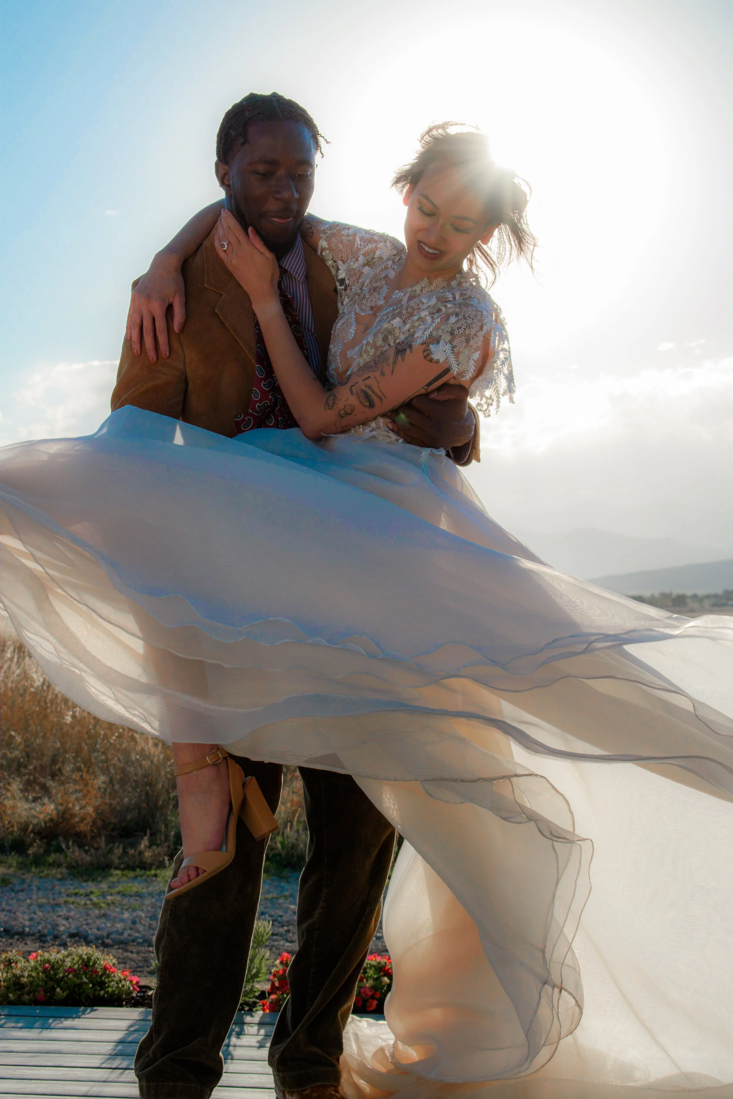 A couple posing outdoors, with the man holding the woman in a wedding dress, backlit by the sun.
