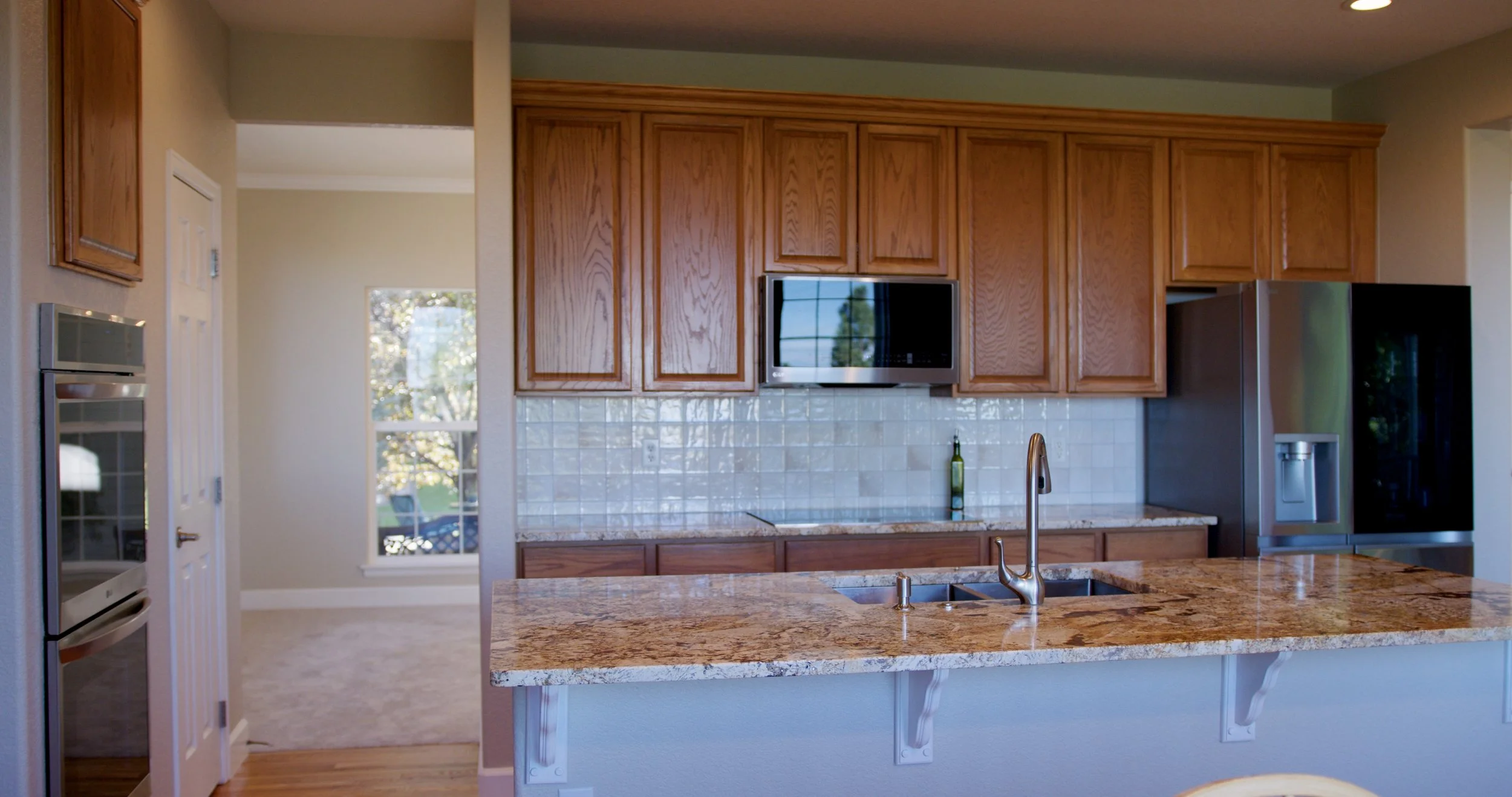 Kitchen with wooden cabinets, granite countertop island, stainless steel appliances including a microwave and refrigerator, tiled backsplash, and a view into a dining area with a window.