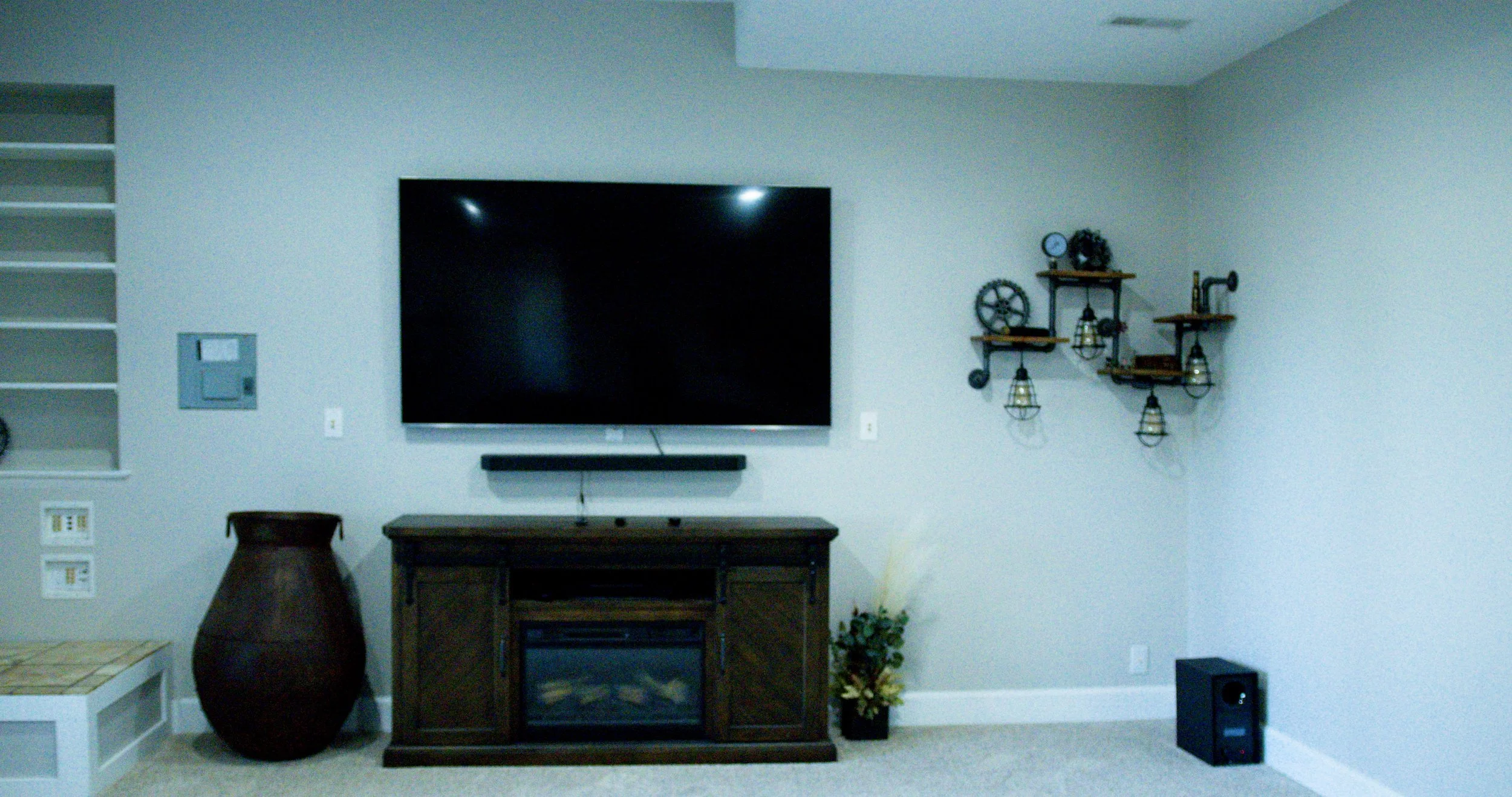 Living room with a large flat-screen TV mounted on a light-colored wall, a wooden console with a fireplace insert, a large brown vase, a floral arrangement in a black pot, and decorative wall shelves with small industrial-style lanterns.