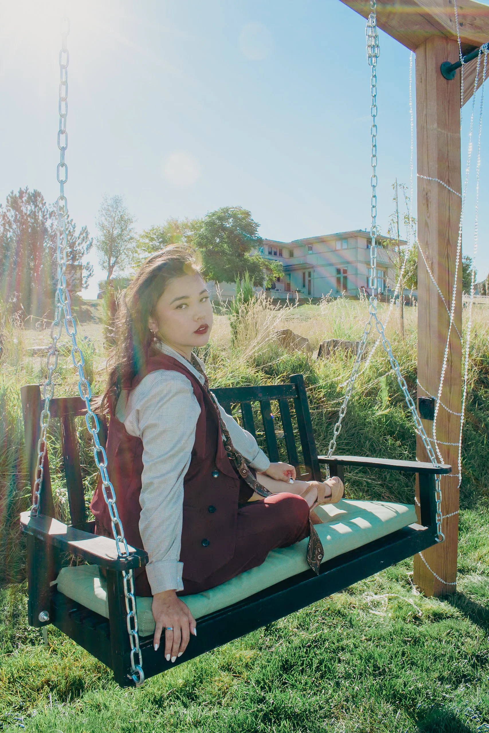 A woman with dark hair and red lipstick sitting on a black wooden porch swing with white cushion, outdoors with sunshine, grass, trees, and a house in the background.