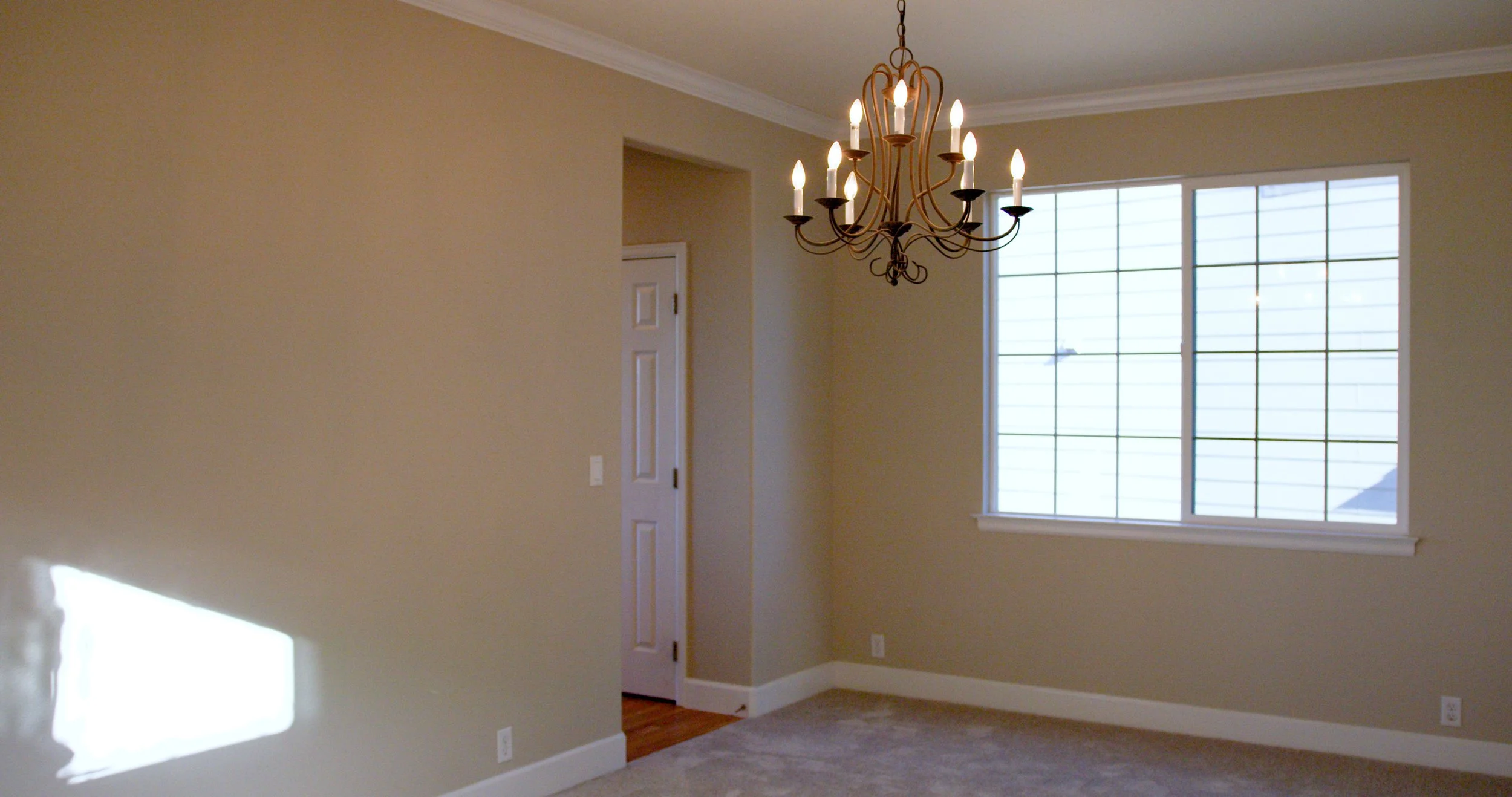 Empty room with a large window, chandelier, beige walls, and carpeted floor.