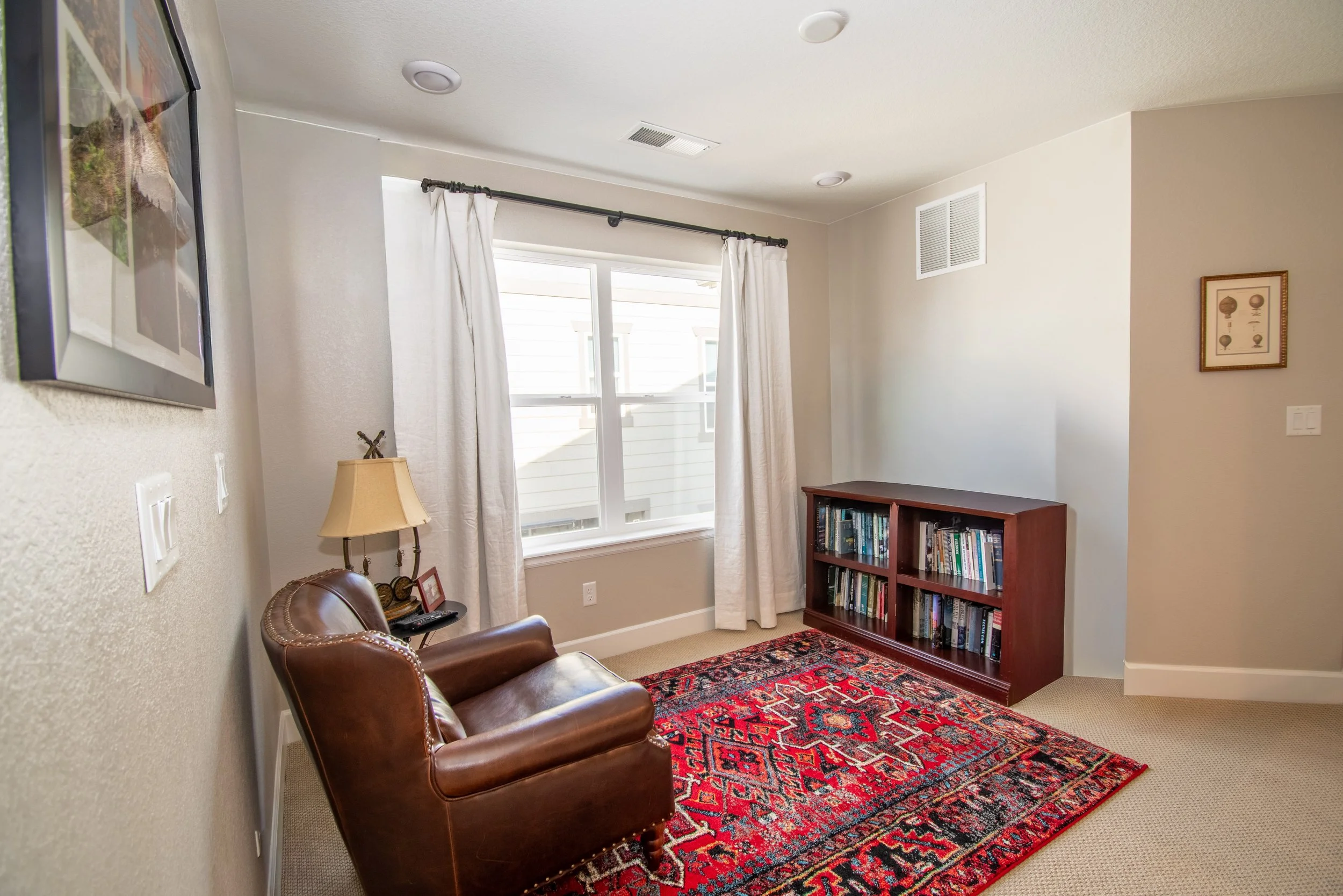 A cozy living room with a brown leather armchair, a small side table with a lamp and framed photo, a window with white curtains, a red patterned rug, and a bookshelf filled with books, beige walls, and a framed picture on the wall.