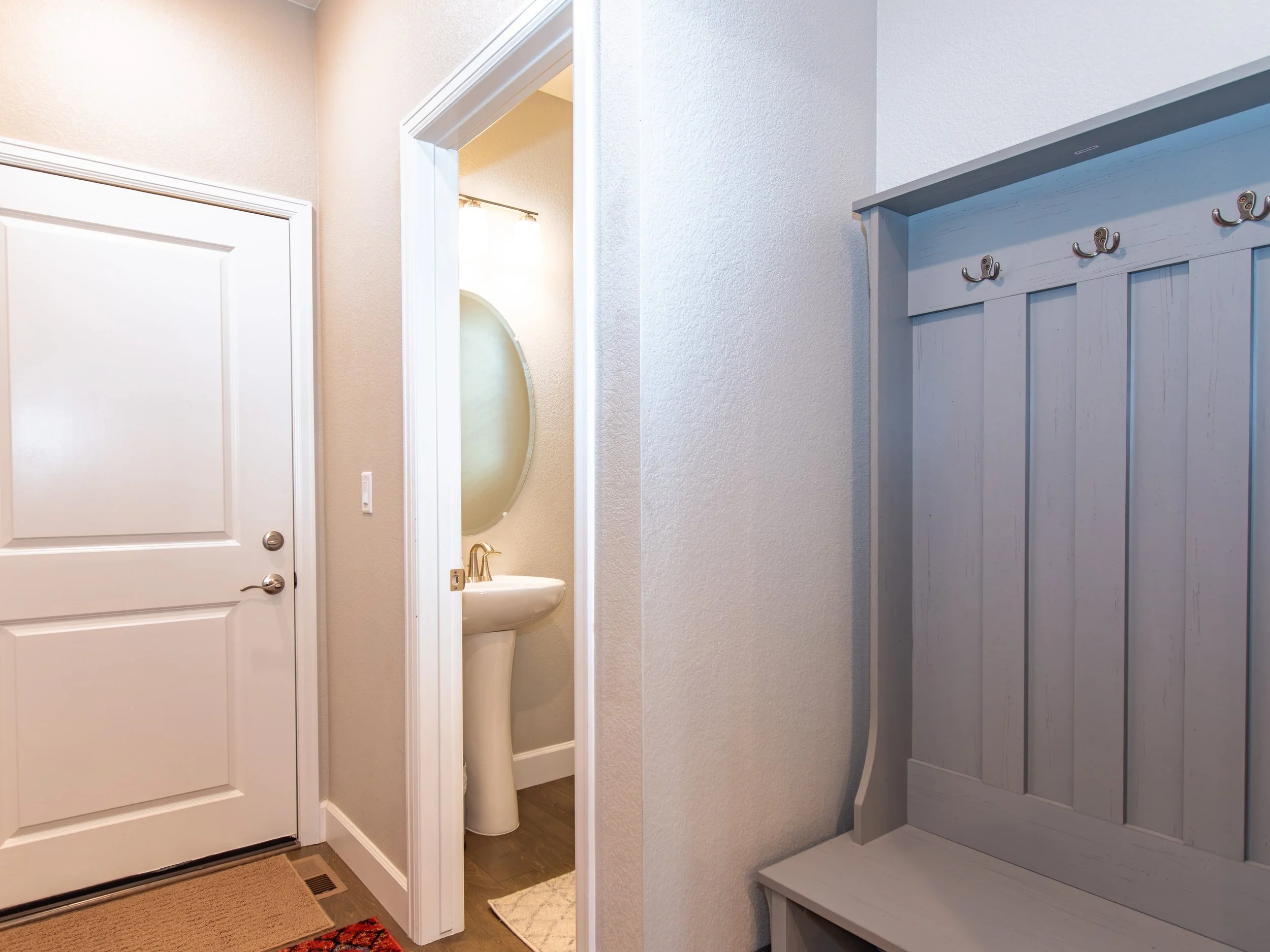 Entryway with a white door, a hallway leading to a bathroom with a pedestal sink and oval mirror, and a grey wooden coat rack with hooks.