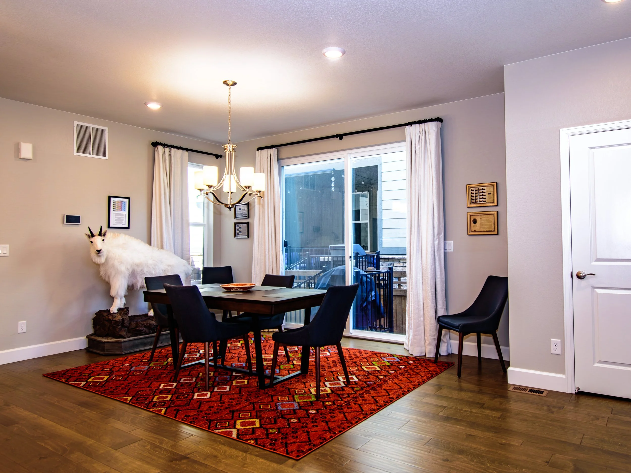 A dining room with a dark wood table and four black chairs, a red patterned rug underneath, a white animal mounted on a stand at the back wall, a sliding glass door with beige curtains, a chandelier hanging above the table, and two chairs at the wall