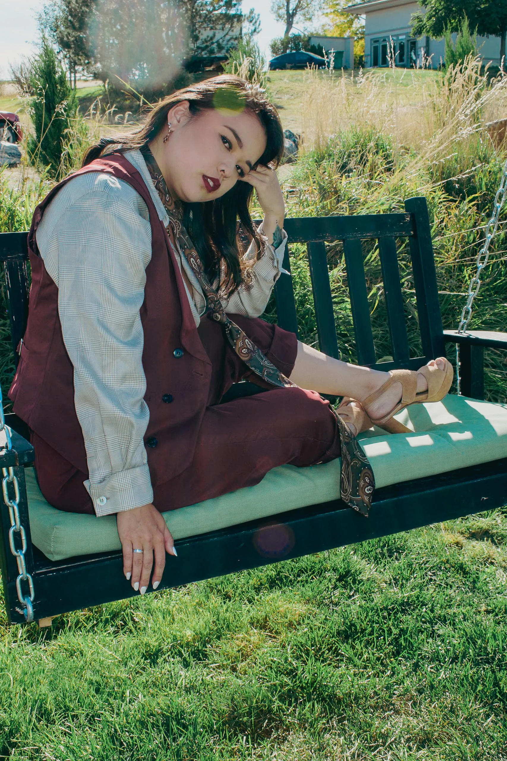 A woman sitting on a swing bench outdoors, with greenery and a house in the background. She is dressed in a beige shirt, maroon vest and pants, with high-heeled shoes, and has dark hair and makeup.