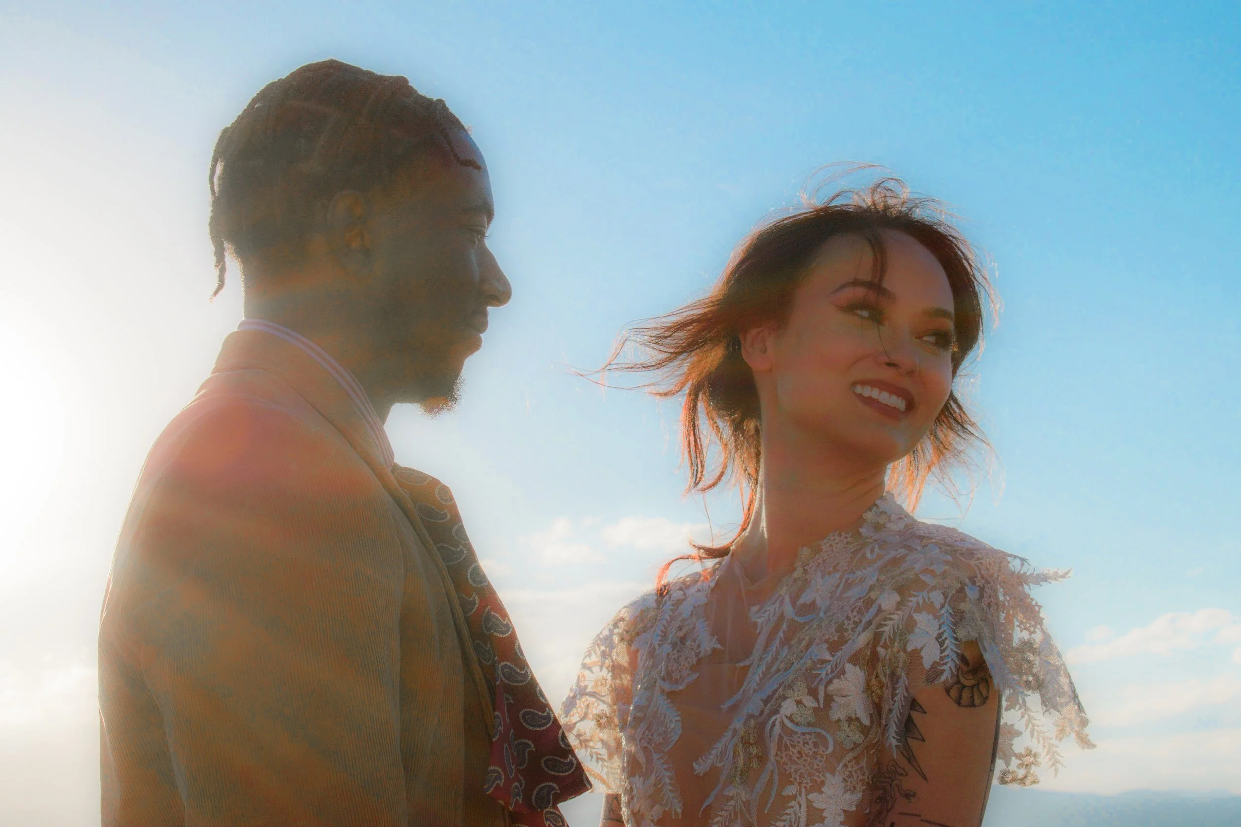 A man and woman standing outdoors against a blue sky with clouds. The man is in profile, wearing a tan suit with a patterned tie. The woman is smiling, looking away, with wind-blown hair and wearing a lace dress.