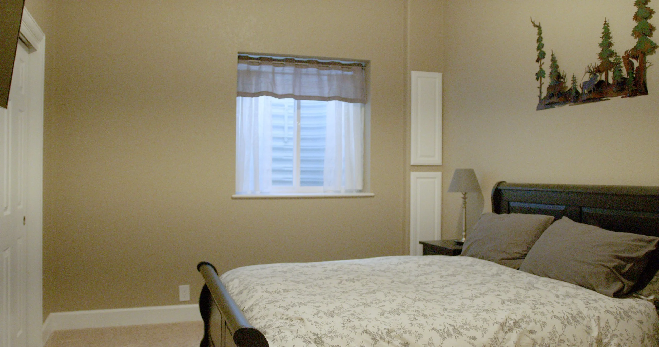A bedroom with beige walls, a window with sheer curtains, a black bed with floral bedding, gray pillows, a nightstand with a lamp, and a landscape painting of trees on the wall.