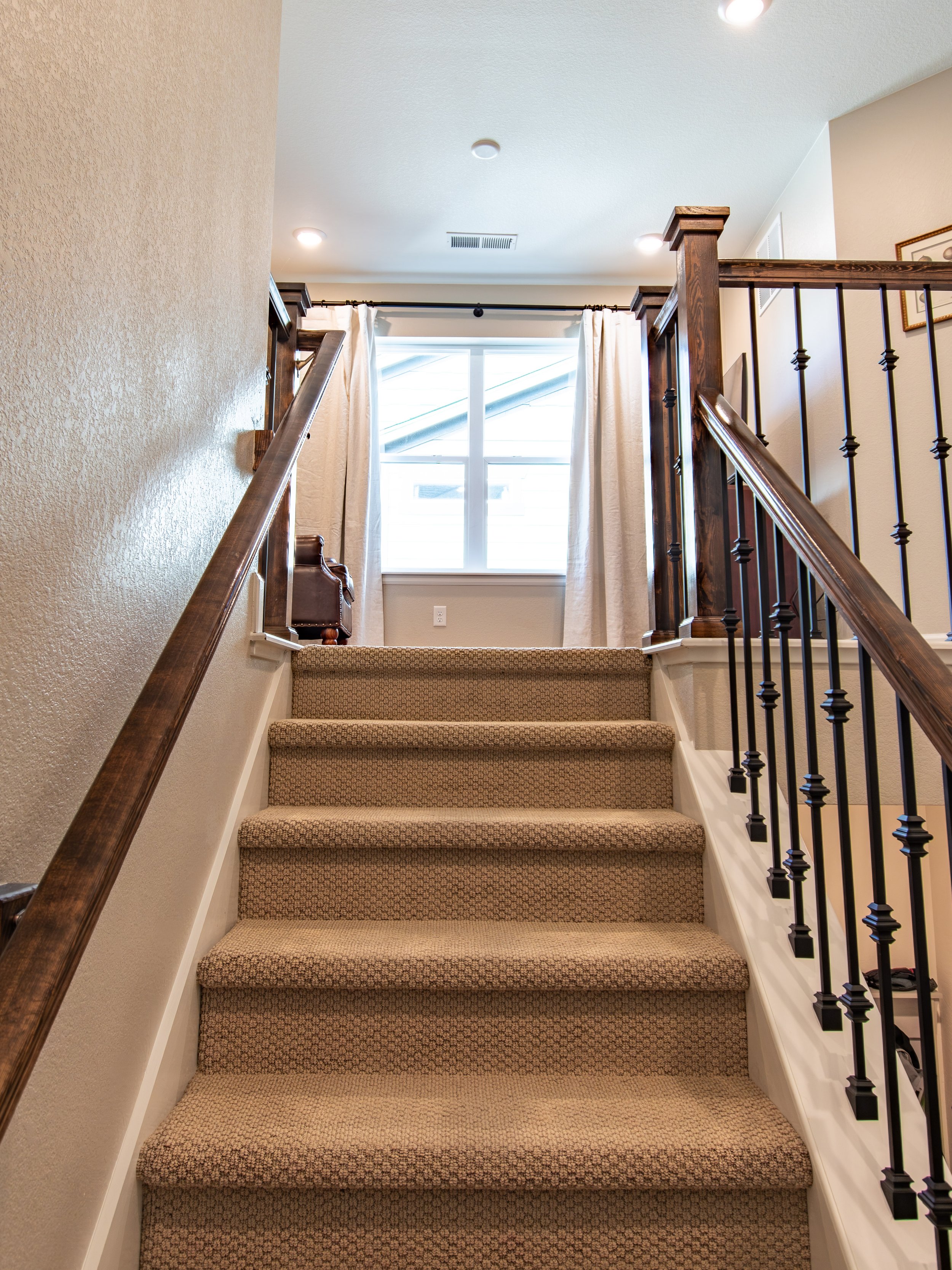 Carpeted staircase leading up to a bright room with window, beige curtains, and wooden handrails on both sides.