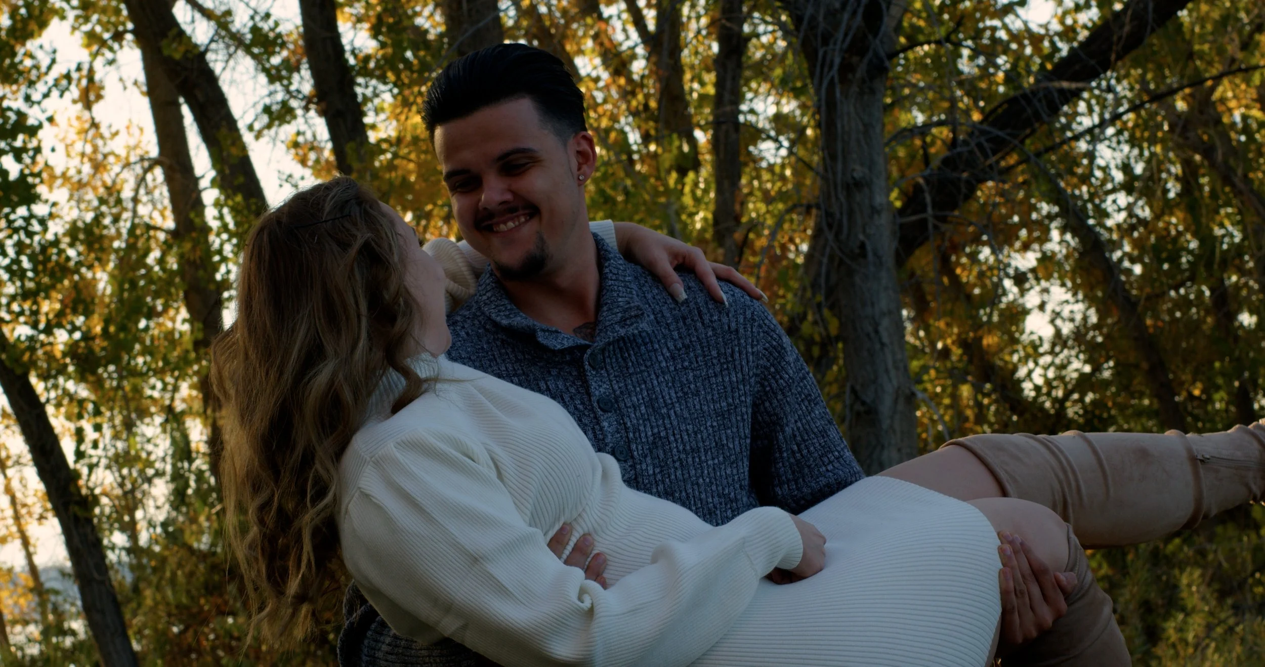 A smiling man is holding a woman in his arms outdoors during fall, with yellow and orange leaves on trees in the background.