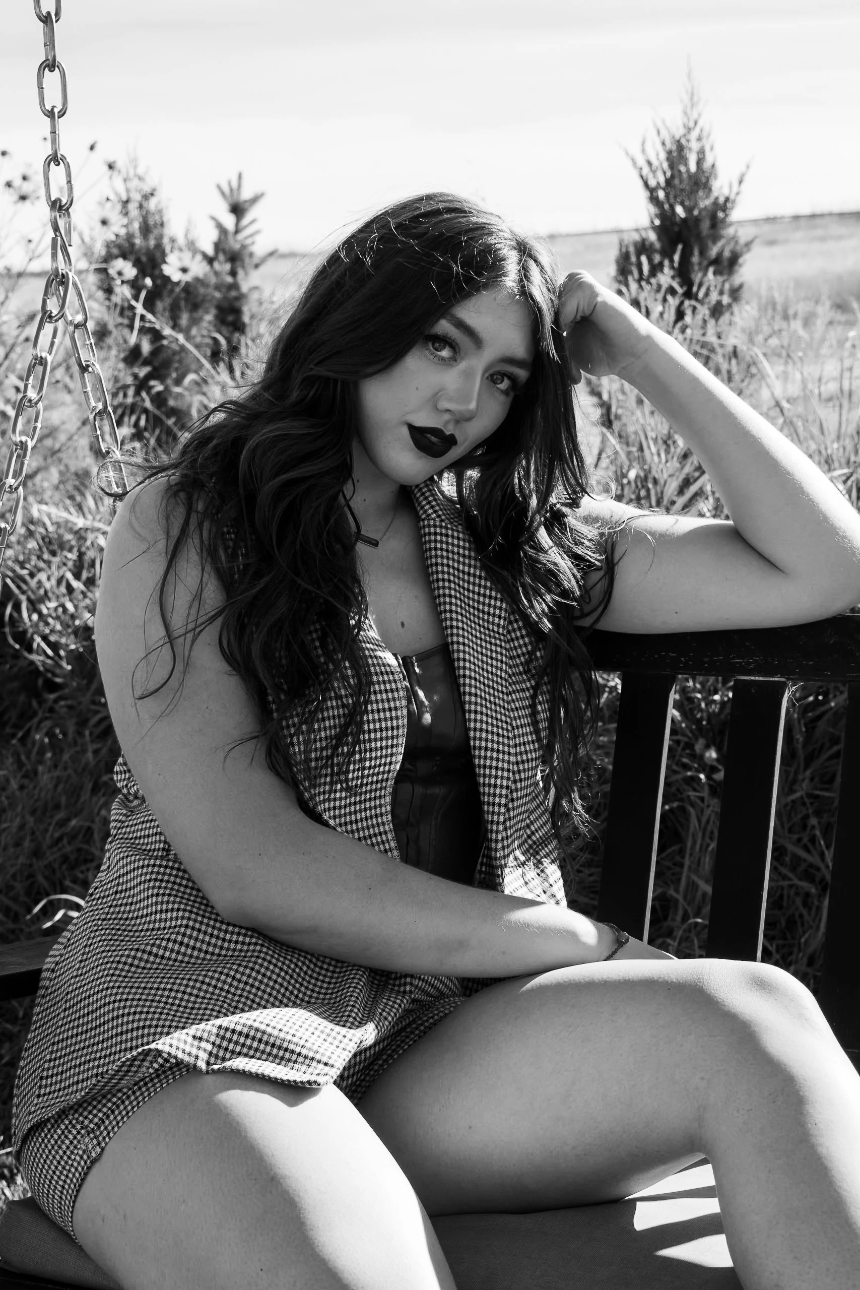 A young woman with long dark hair sitting outdoors on a swing, resting her arm on a bench, looking at the camera with a slight smile, in black and white.