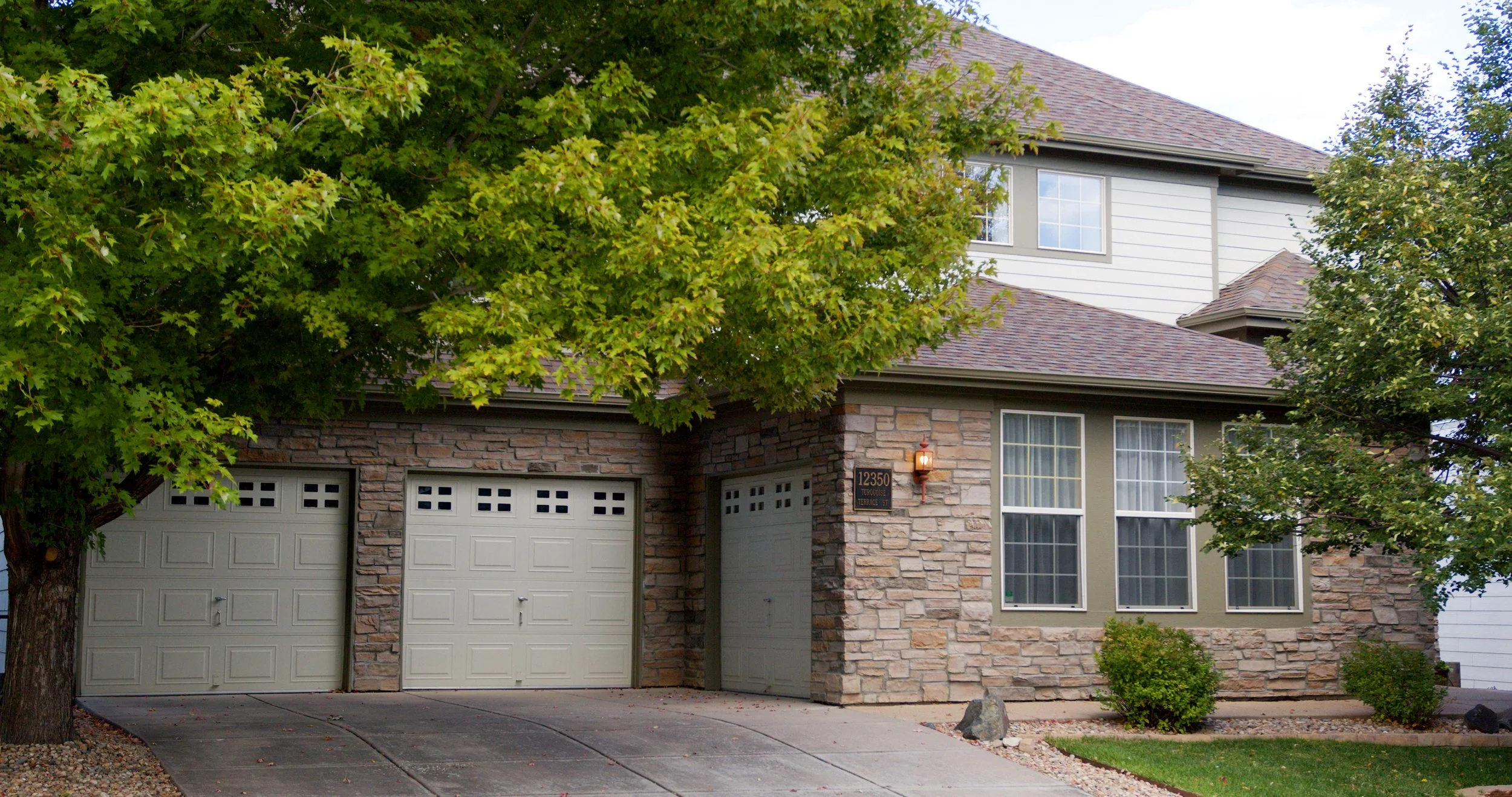 Front of a house with two beige garage doors, stone exterior walls, three large windows, green trees, shrubs, and a landscaped yard.