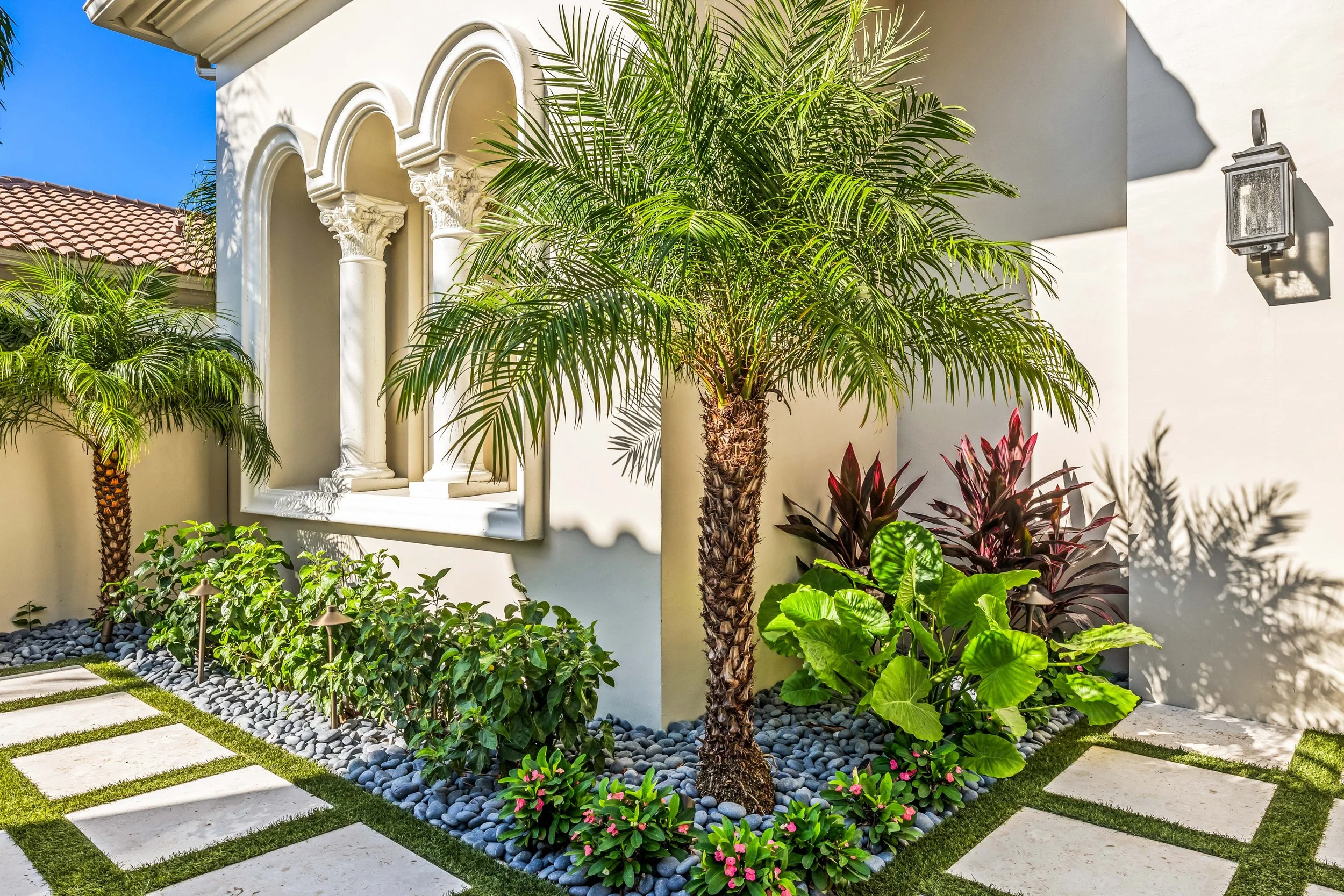 Luxury portico entry with sculpted tree, layered tropical foliage, and architectural lighting.