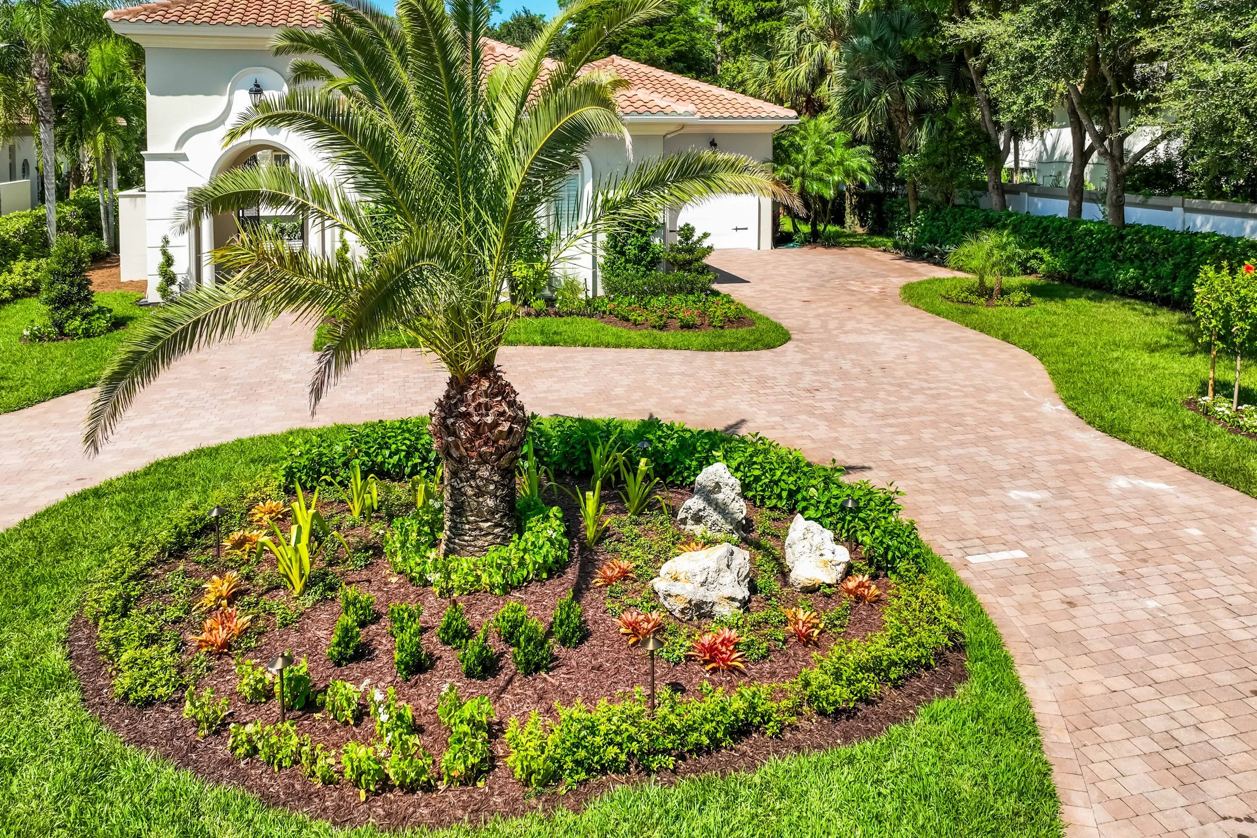 Circular landscape bed with foxtail palm, tropical plantings, decorative boulders, and a paver driveway at a Southwest Florida home.
