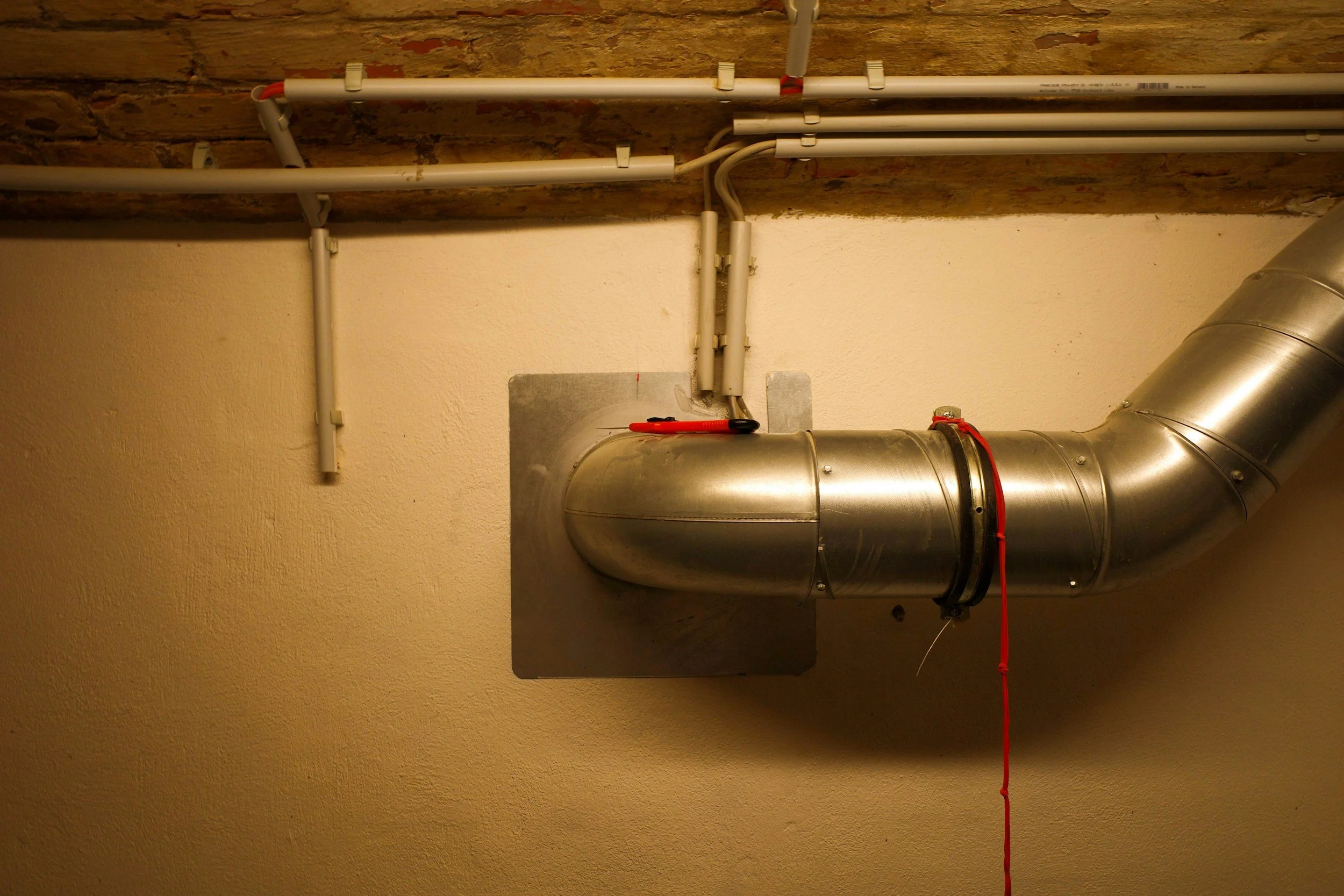 Metal ventilation duct pipe attached to the wall with a red string hanging from it, situated below exposed brick ceiling and above a beige wall.