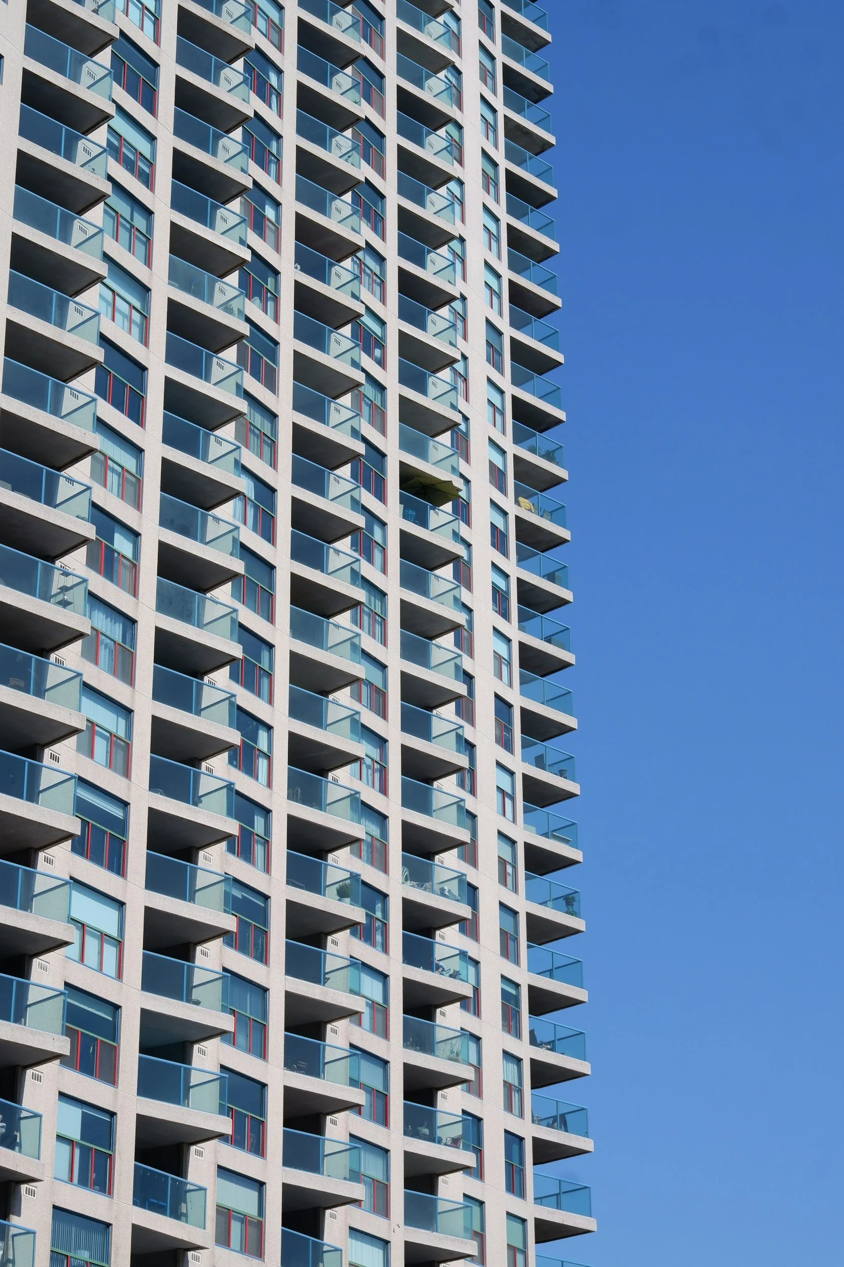 A tall modern apartment building with multiple floors, featuring balconies with glass railings, many windows, and a clear blue sky in the background.