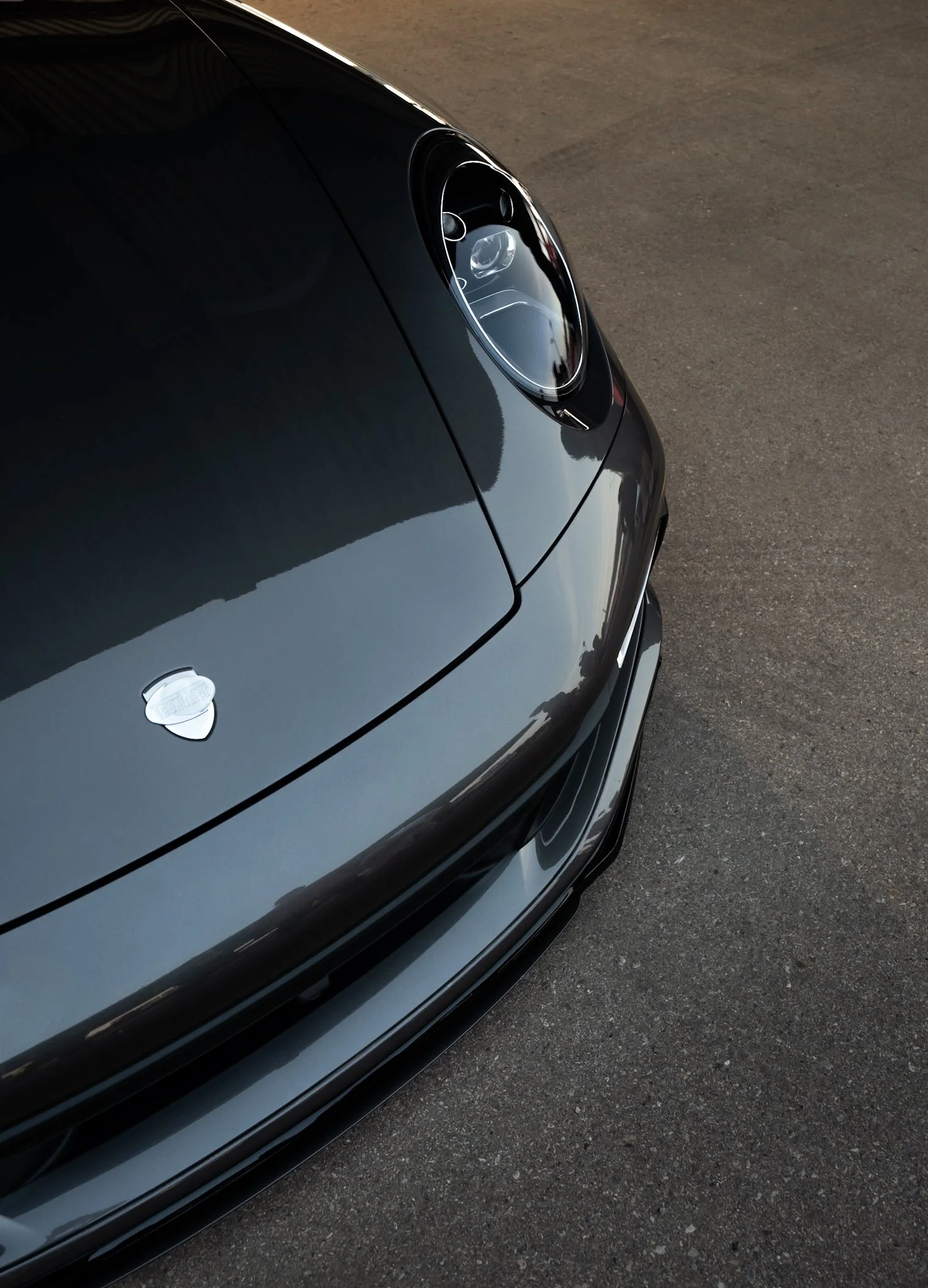 Close-up of the front part of a sleek, dark-colored sports car, including the headlight and part of the hood, parked on an asphalt surface.