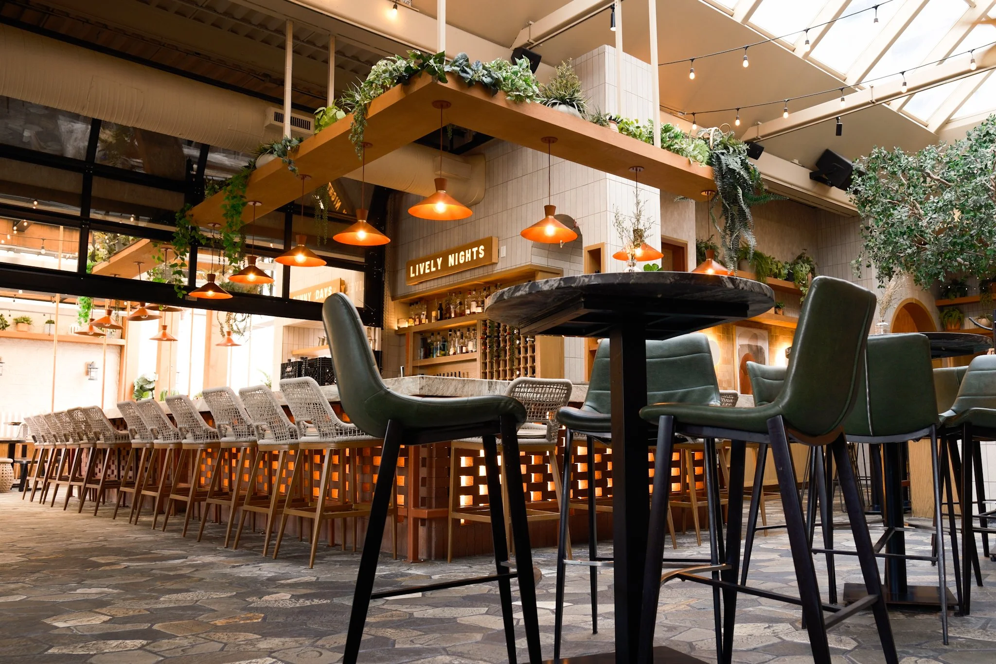 Interior of a modern restaurant with a high ceiling, hanging orange lamps, a bar area, and a variety of plants on upper shelves, with a mix of chairs at tables.