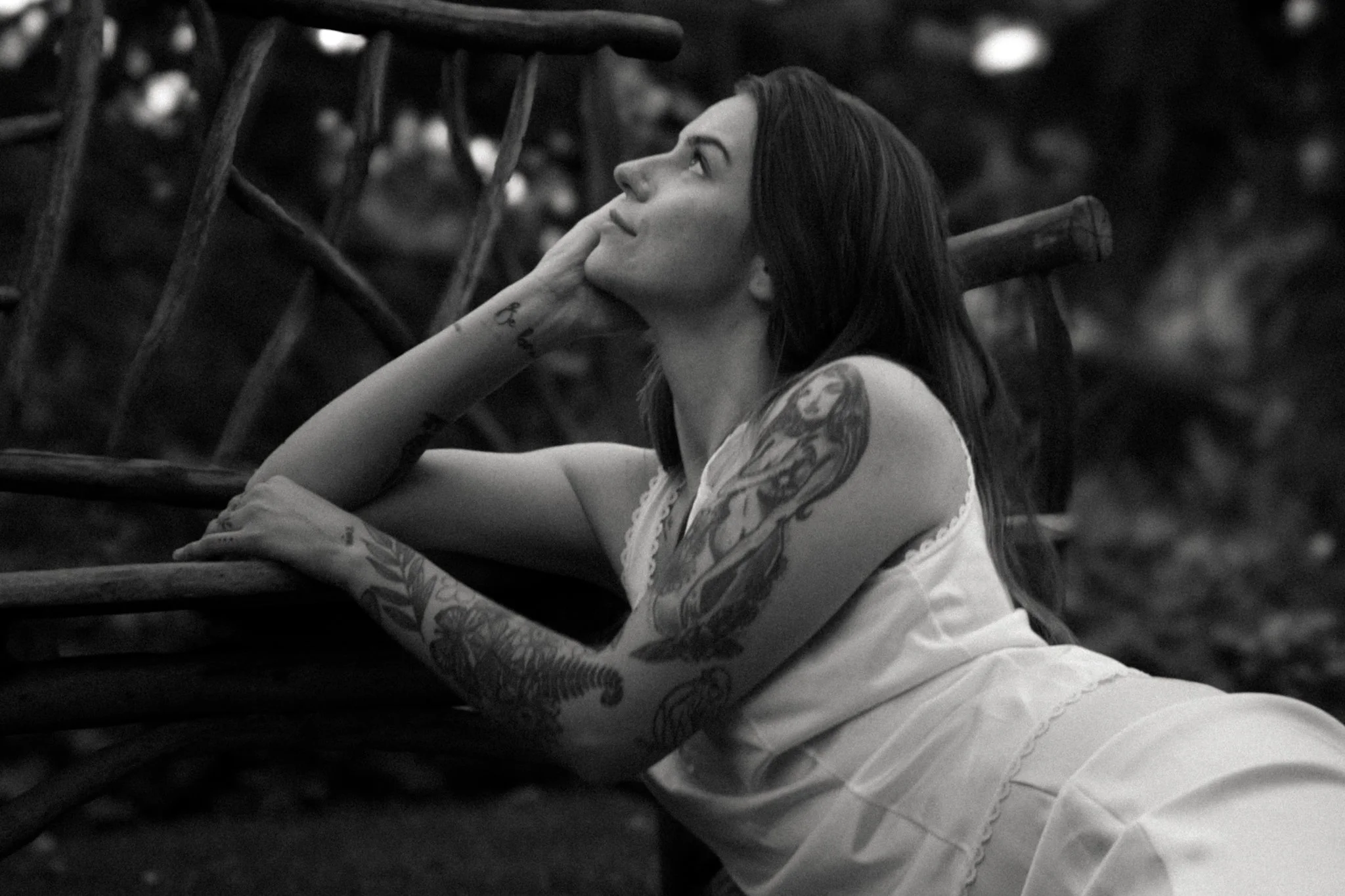 A woman with tattoos on her arm, dyed black hair, and dressed in a white top and light-colored skirt, is sitting outdoors, resting her chin on her hand, and looking up thoughtfully. There are wooden chairs and a blurred background of trees or bushes.