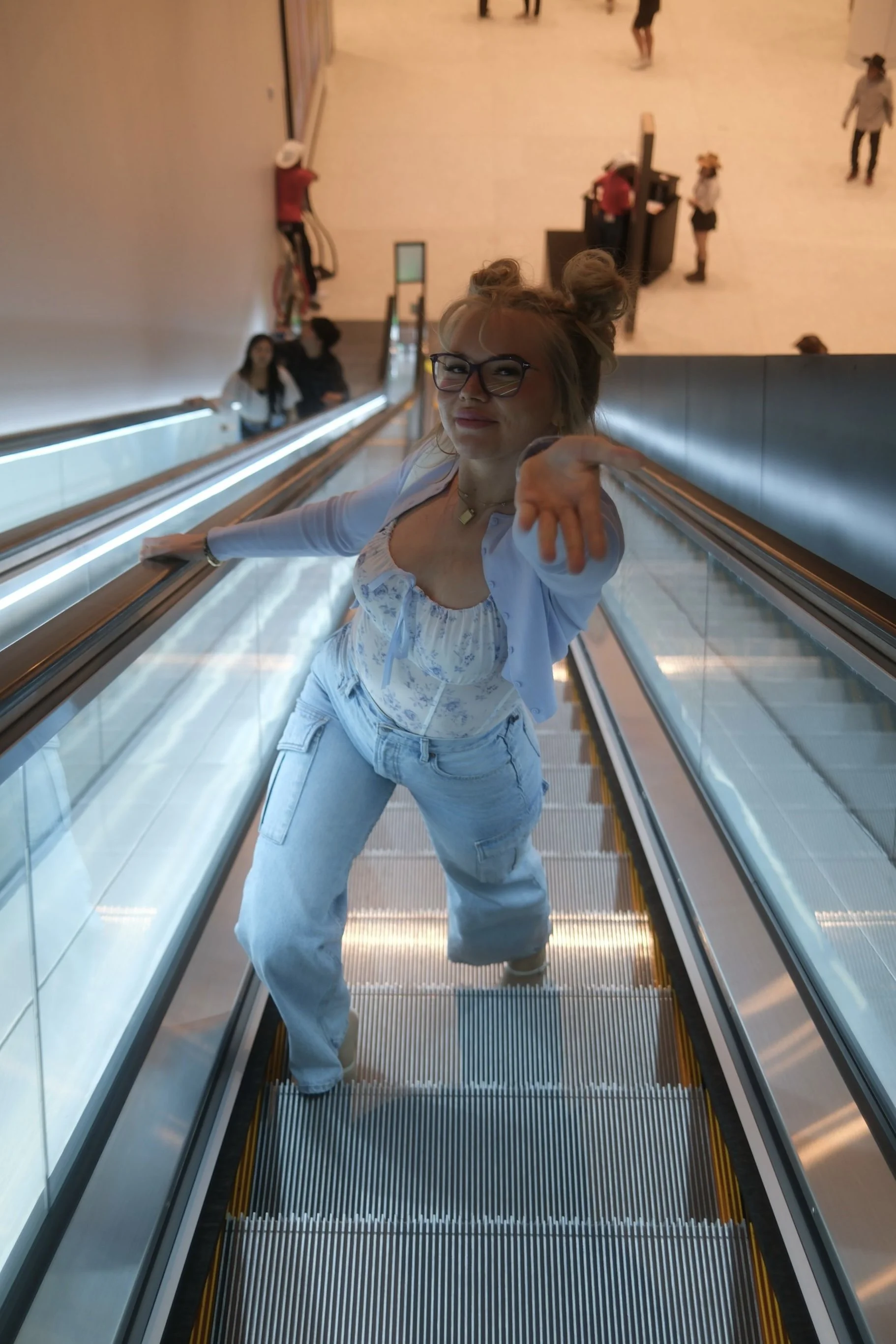 A woman with glasses and blonde hair styled in buns is riding an escalator, smiling and pointing at the camera, in calgary bmo centre.