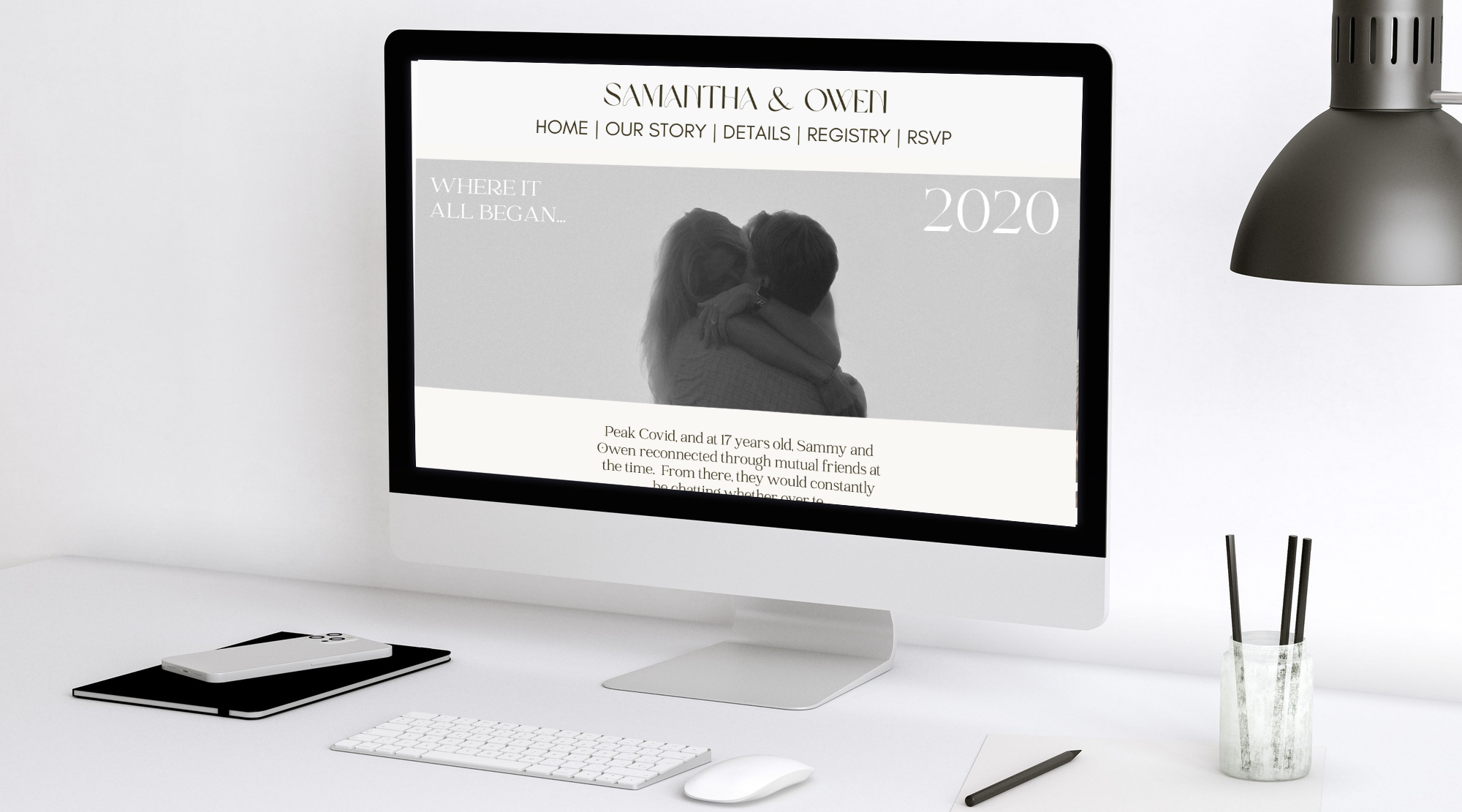 Computer monitor displaying a wedding website for Samantha & Owen with a black-and-white photo of a couple hugging, on a white desk with a keyboard, mouse, notebook, pen, and a lamp.