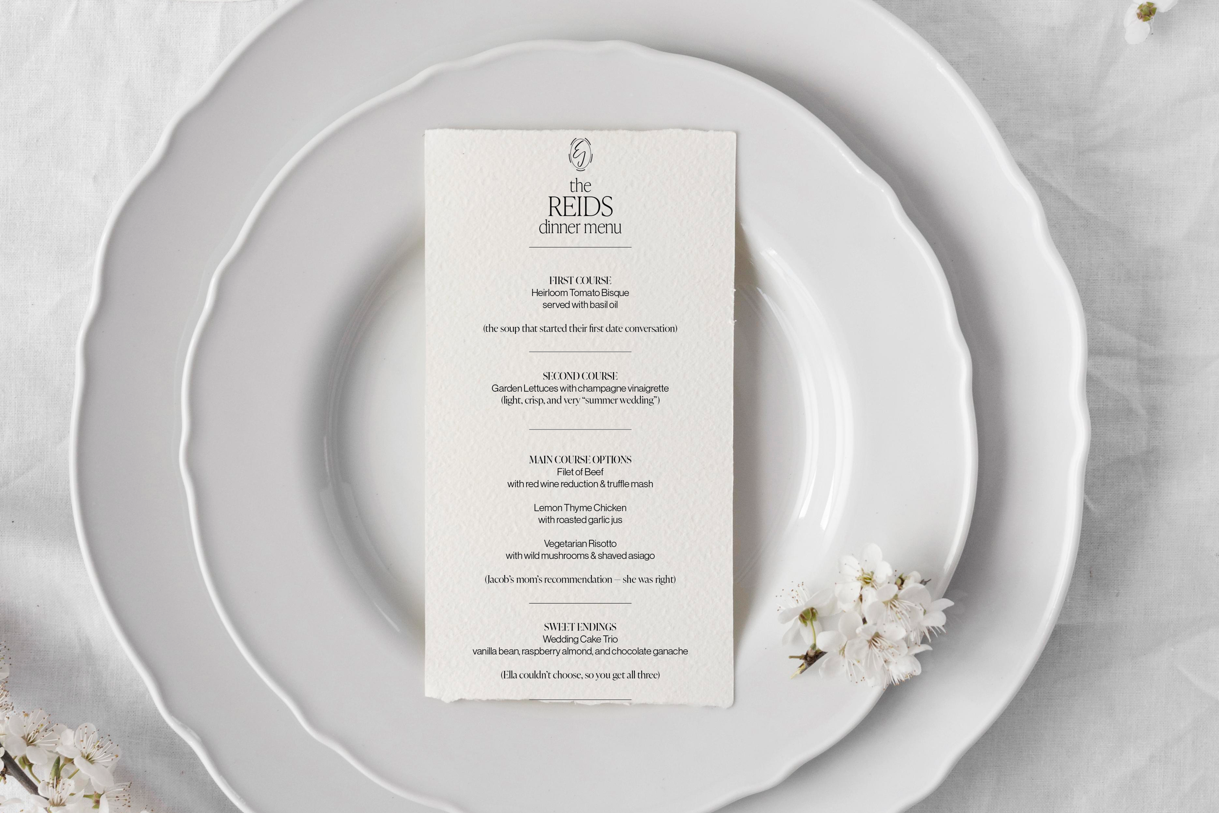 A white dinner plate with scalloped edges, layered on top of a larger matching plate, with a white textured paper menu placed in the center. The menu is titled 'The Reids Dinner Menu' and lists various courses. Small white flowers are placed around t