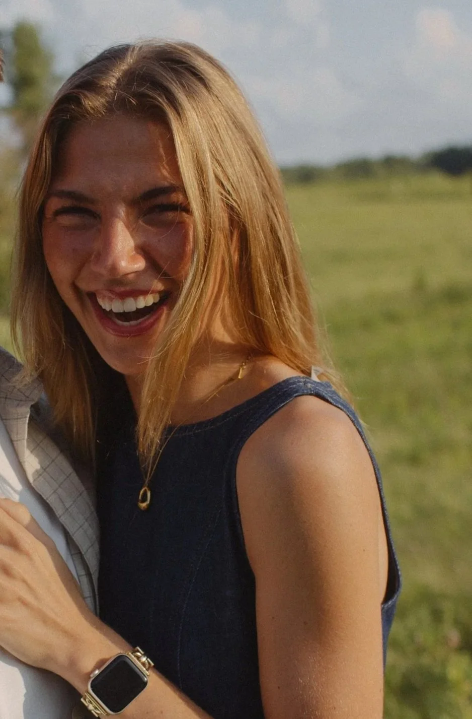 A woman smiling outdoors on a sunny day, wearing a sleeveless dark denim top and a watch on her wrist.