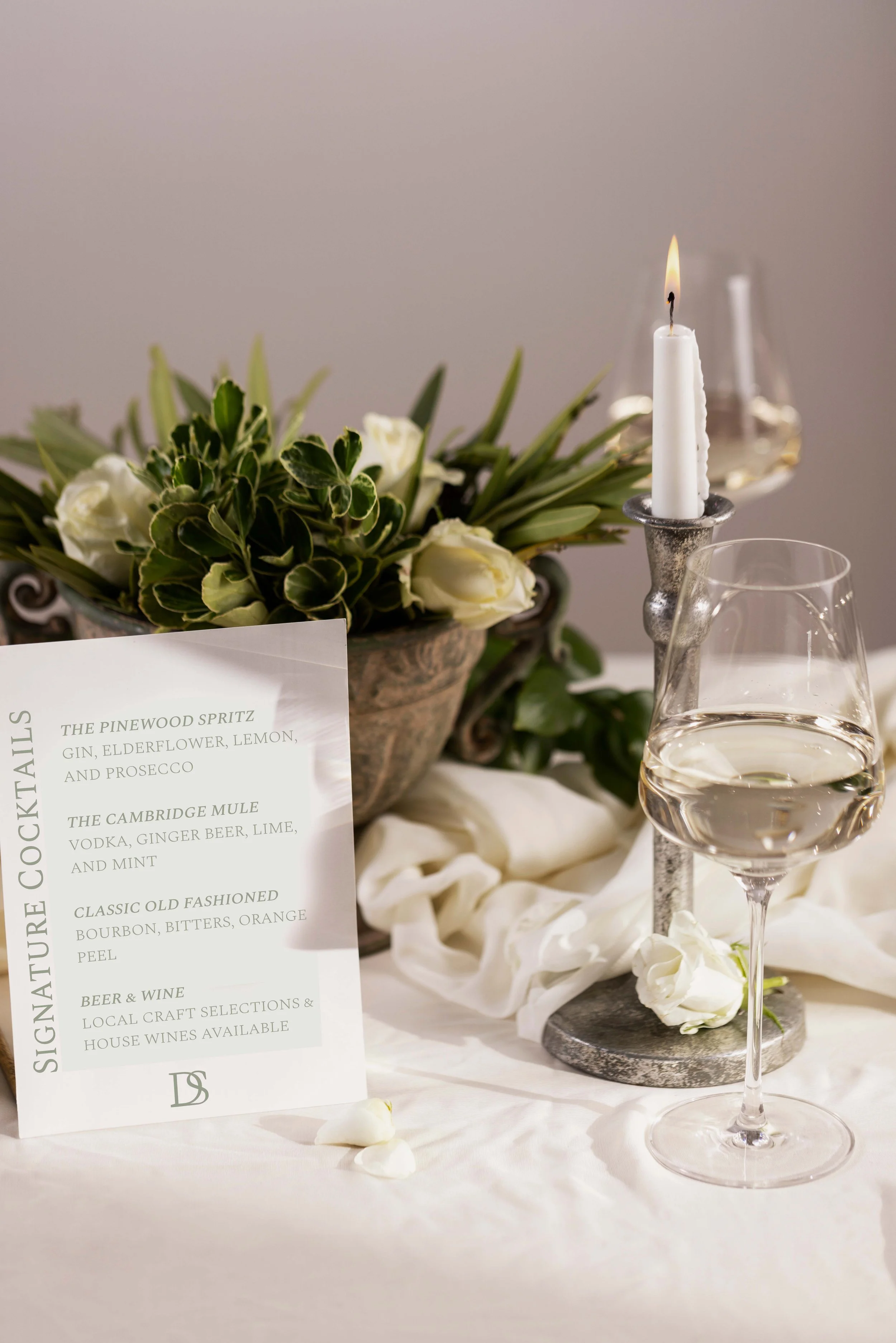 A table centerpiece with a lit white candle in a candlestick, a glass of white wine, a bouquet of white roses and greenery, and a sign listing signature cocktails at an event or wedding.