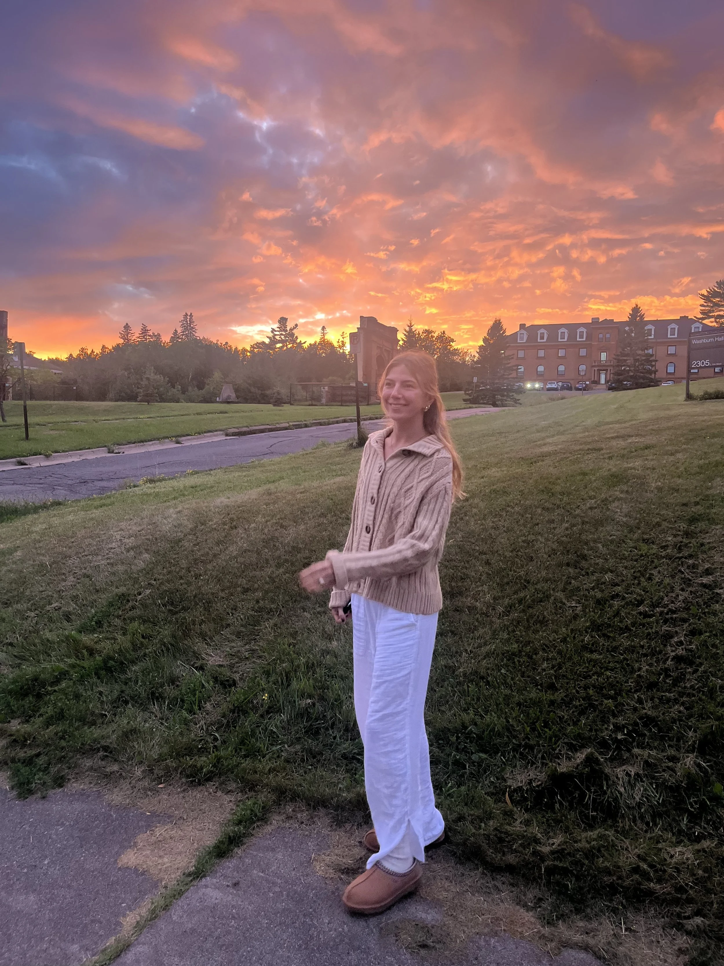 A young woman with long red hair wearing a beige sweater, white pants, and tan shoes, standing outdoors on grass during sunset with colorful clouds in the sky, smiling at the camera.