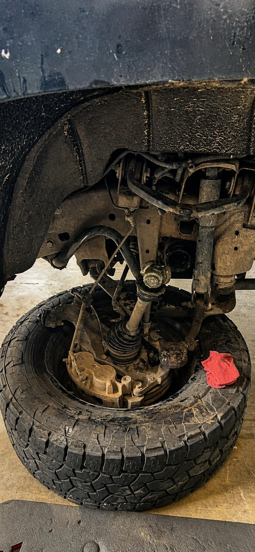 A vehicle's front wheel removed, exposing suspension components, steering linkage, and brake system, with a tire resting on the ground and a red cloth nearby.