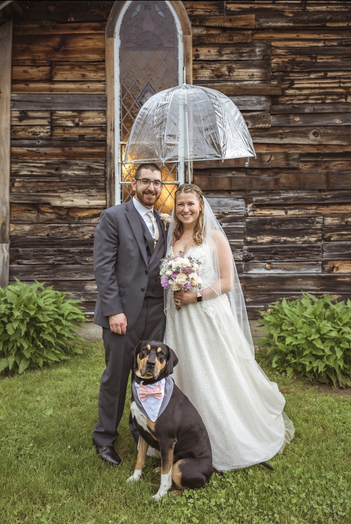 Image of the owner, Karissa, and her husband on their wedding day. Standing in front of a barn with their dog in a tux bandana and holding a clear umbrella to protect from the rain.