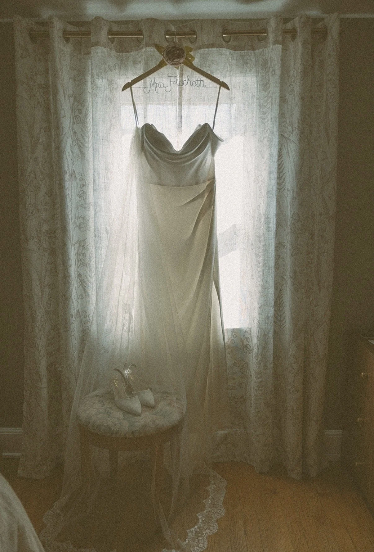 A wedding dress hanging on a hanger attached to a curtain rod in front of a window with sheer curtains. A pair of white bridal shoes are placed on a small cushioned stool underneath the dress.