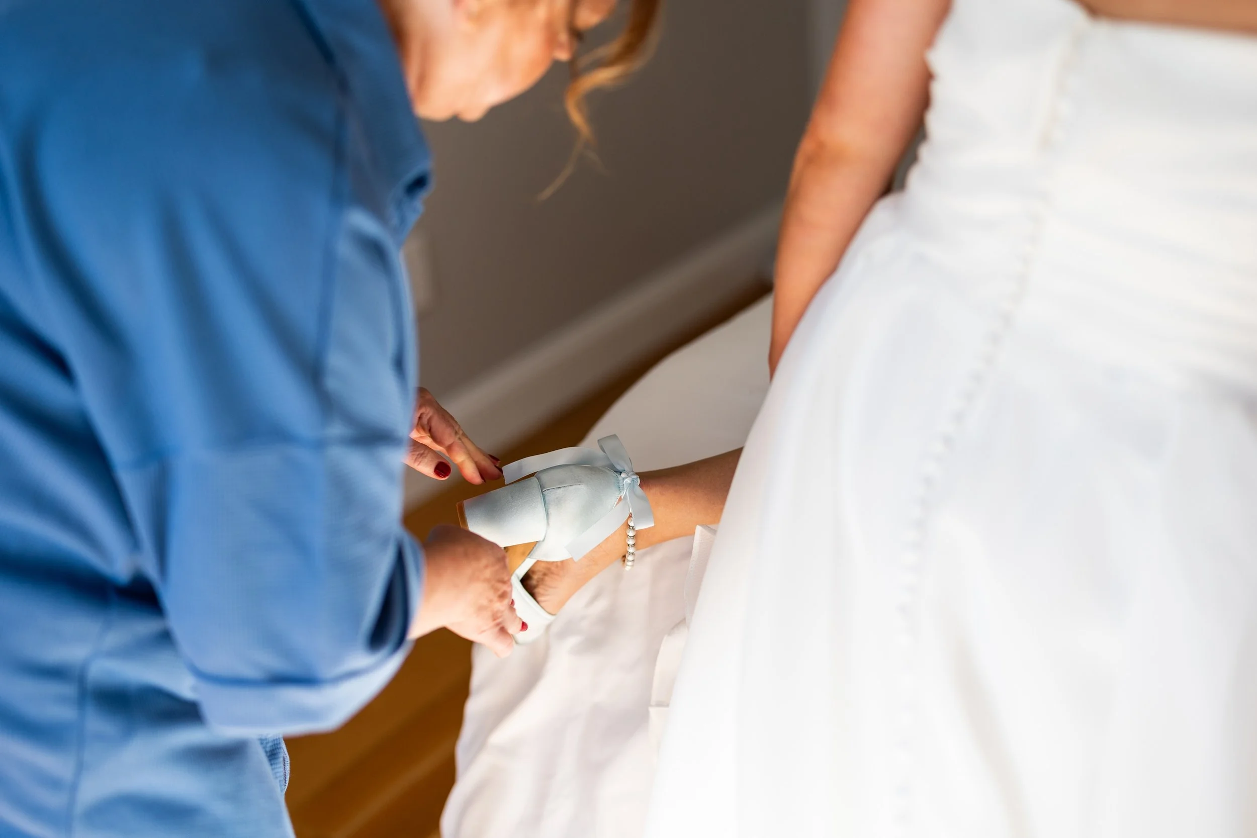 A woman in a blue shirt is helping a bride put on her wedding shoes, which are white with a ribbon and pearl detail.